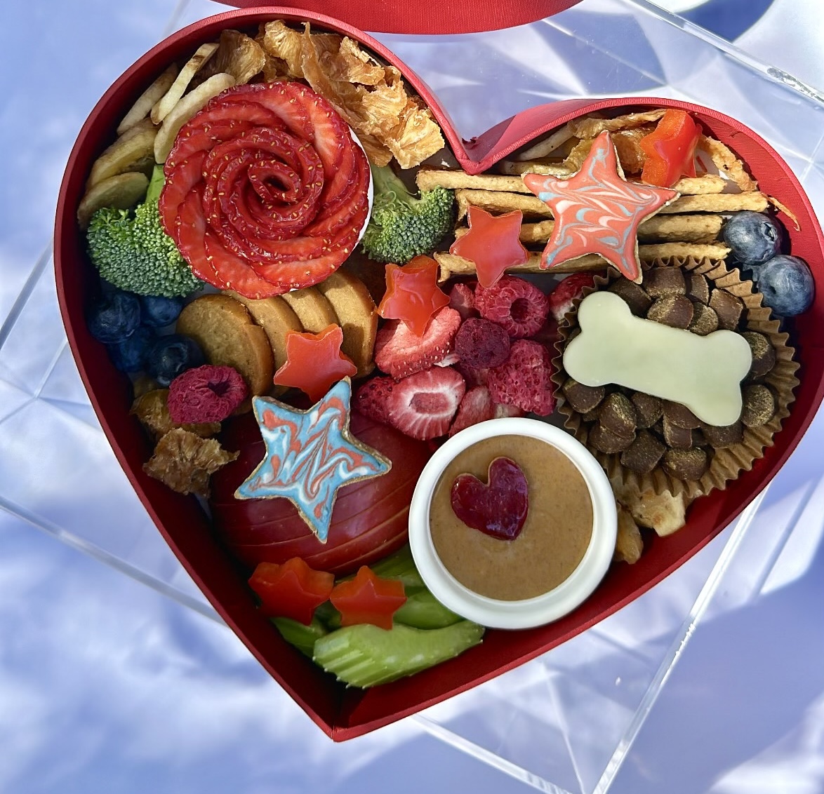 A heart-shaped box filled with a variety of treats including fruit, vegetables, crackers, a peanut butter cup, dog treats shaped like stars and a bone.