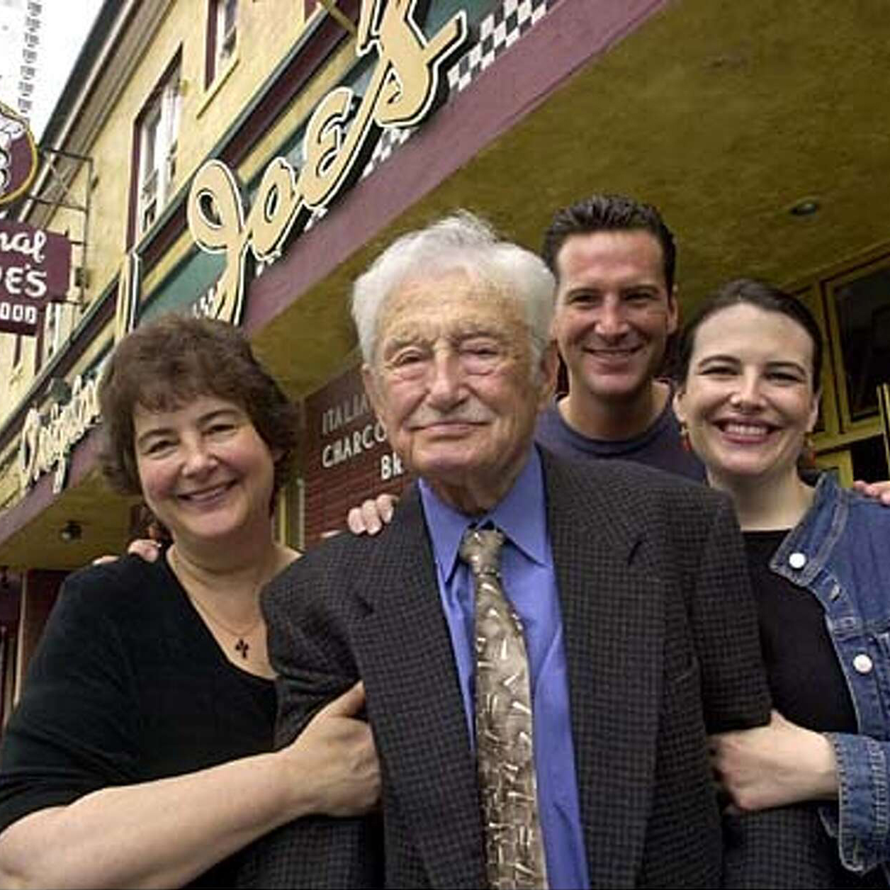 Four people stand closely together, smiling, in front of a restaurant named &quot;Joe's&quot; with vintage-style signage. The elderly man in the front wears a suit.