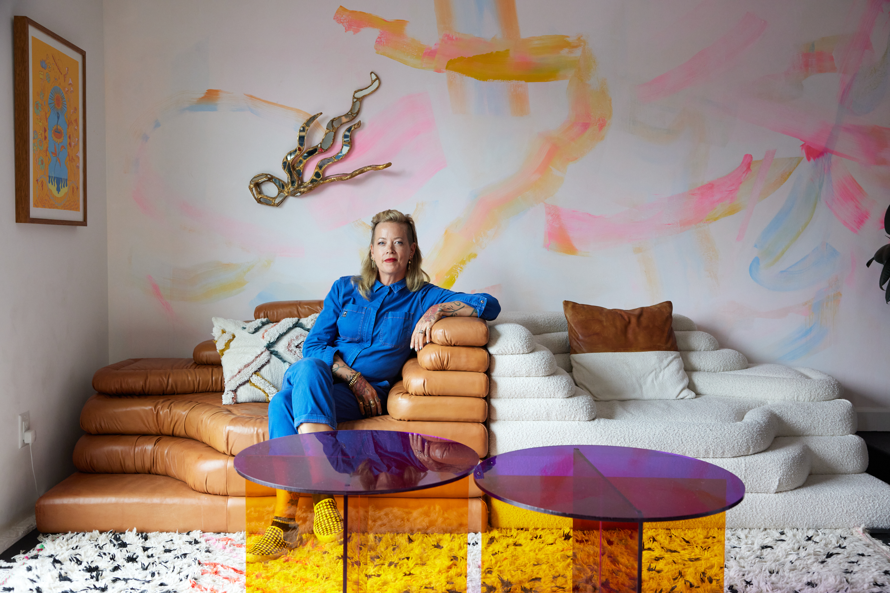 A woman in a blue jumpsuit sits on a tan couch against a pastel-painted wall, beside a white textured couch. Two colorful glass tables are in front of her.