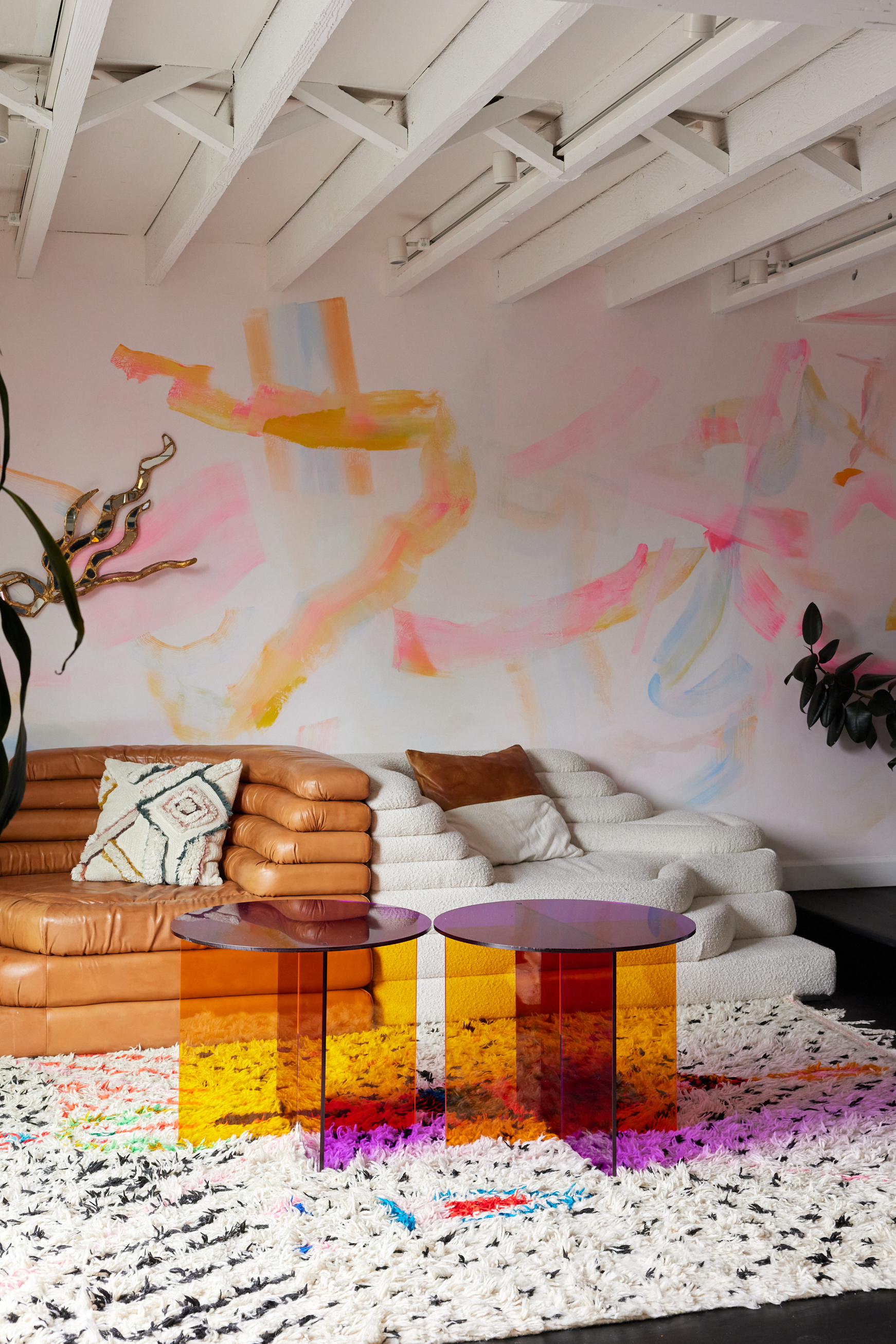 The image shows a colorful, modern living room with tan and white sofas, abstract wall art, a patterned rug, and two small tables with orange and purple translucent bases.