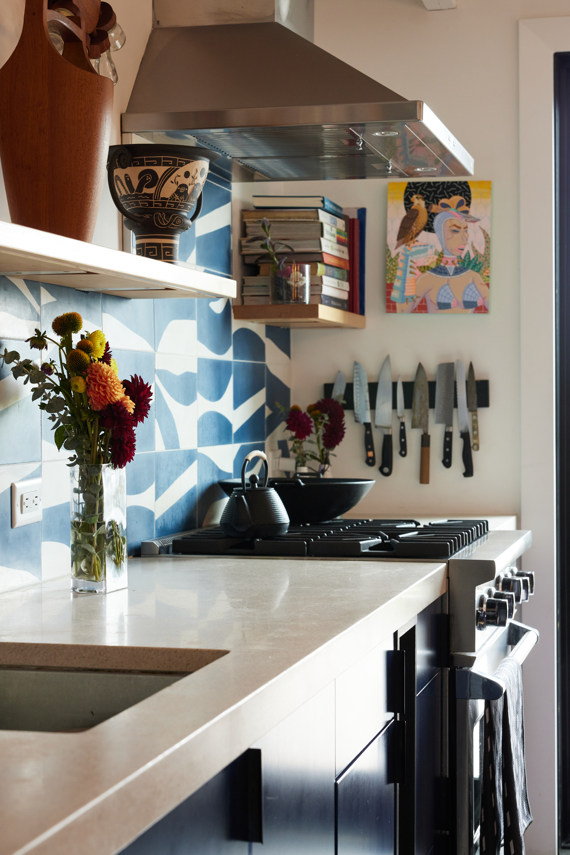 A kitchen with blue and white tiled backsplash, wooden shelves holding books and decor, a knife rack, a stove with a kettle, and fresh flowers in a vase.