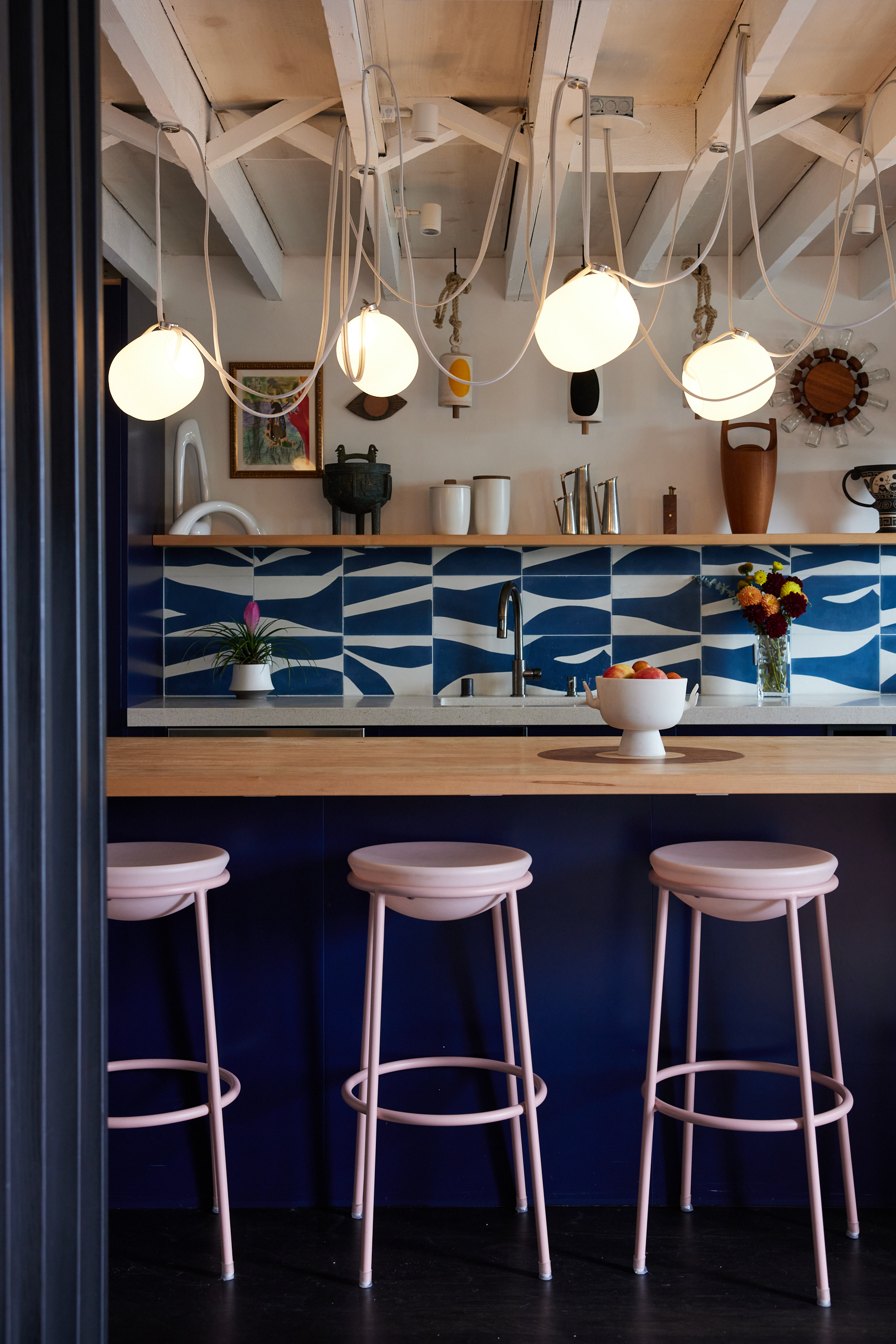 This image shows a stylish kitchen with modern light fixtures, blue patterned backsplash tiles, a wooden countertop, pink stools, and various decor items.