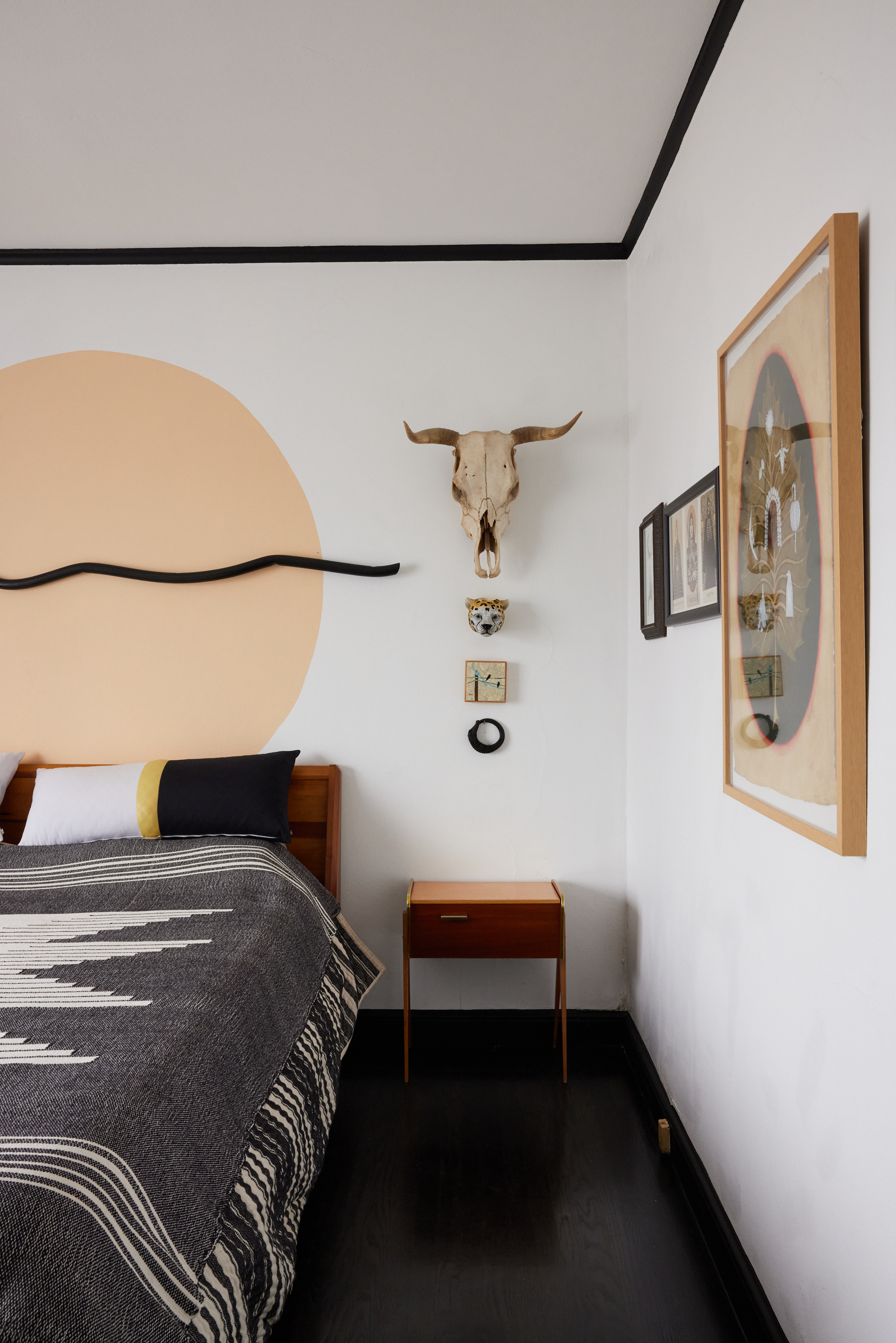 A bedroom features a wooden bed with a patterned blanket, a skull mounted above a nightstand, and framed art. The wall has a large peach circle behind a curving black line.