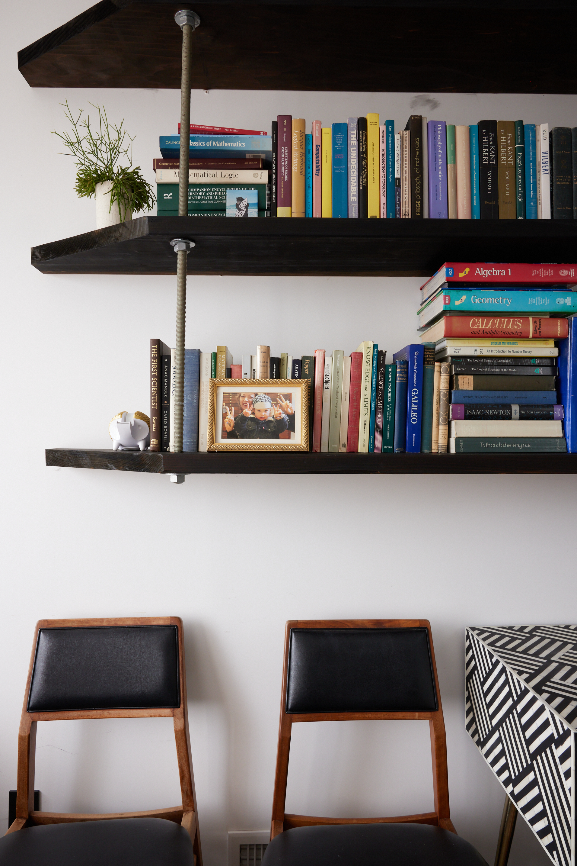 The image shows two wooden chairs with black cushions, a geometric-patterned table, and wall-mounted shelves filled with books, a photo frame, and a plant.