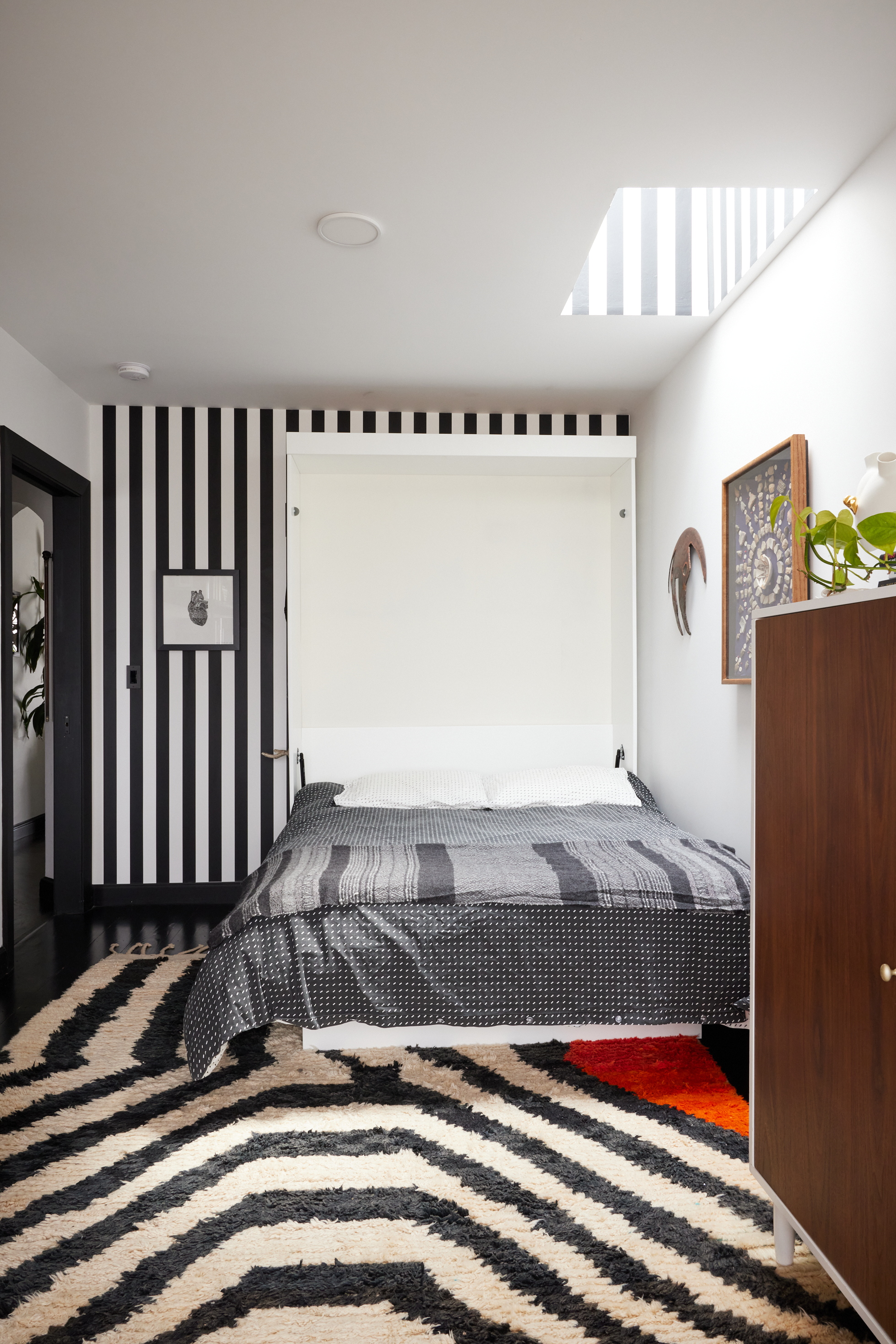 A modern bedroom with black and white striped walls, a bed with a striped gray blanket, and a large chevron-patterned rug. A wooden cabinet and framed pictures adorn the space.