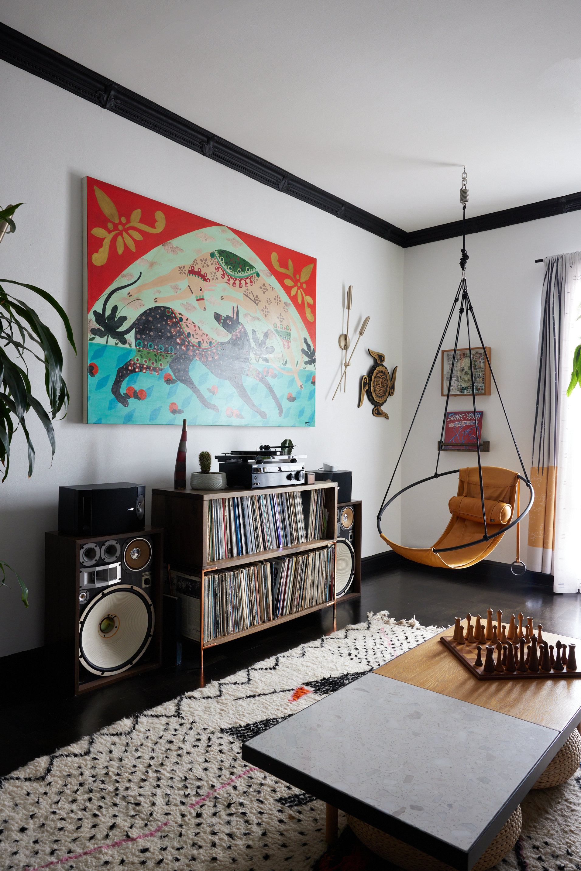 A cozy room with a large, colorful painting of abstract animals above a record player and vinyl collection, speakers, a hanging chair, and a chess set on a coffee table.
