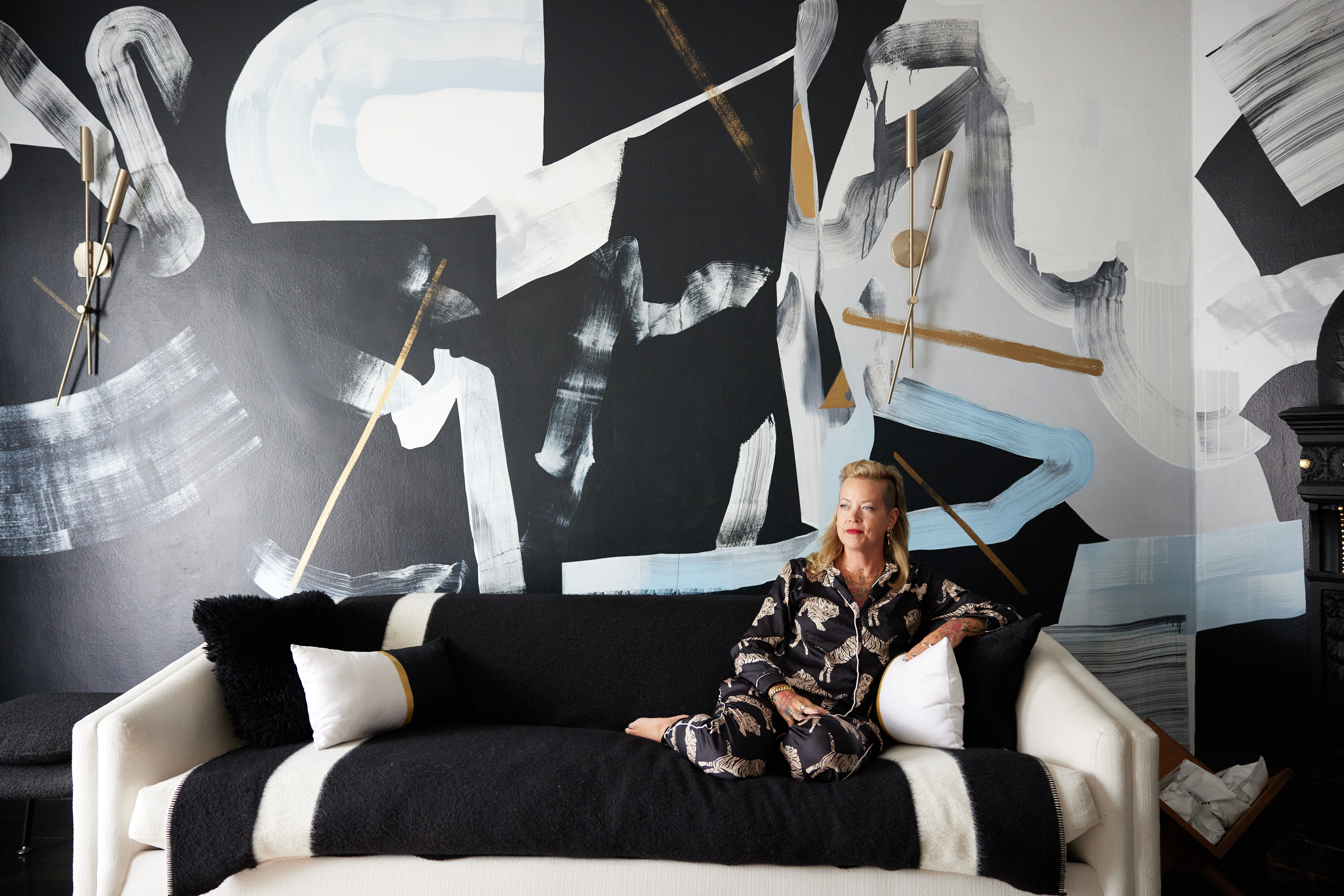 A woman in patterned clothing sits on a modern black-and-white sofa against a dramatic abstract mural with bold brushstrokes and geometric shapes.