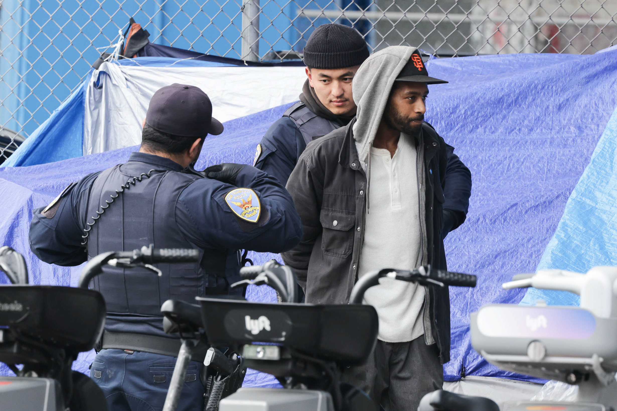 Two police officers detain a man in front of a chain-link fence covered with a blue tarp. Several Lyft rental bikes are in the foreground.