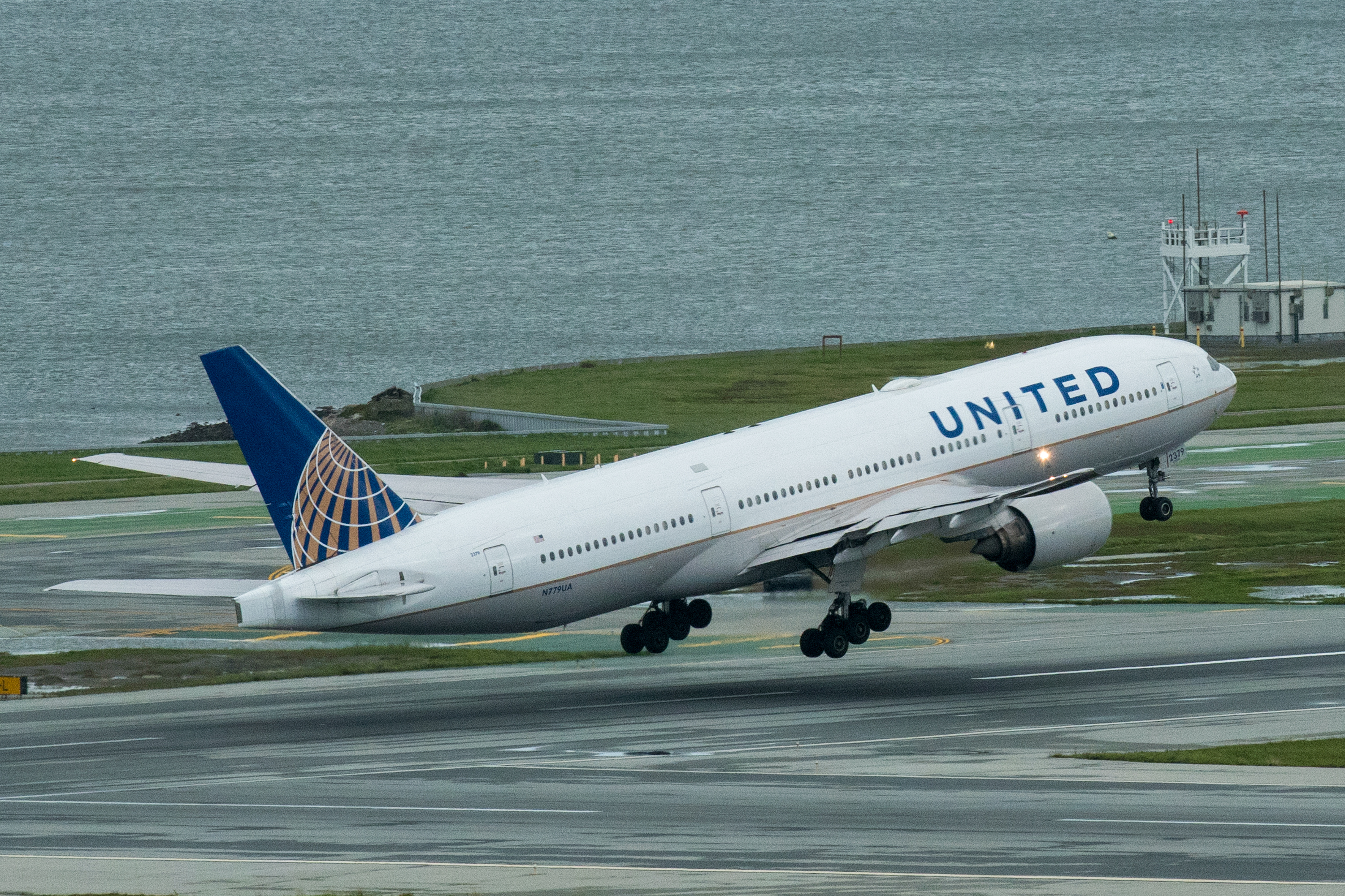 A white airplane with "United" on the side is taking off from an airport runway, with the ocean visible in the background on a cloudy day.