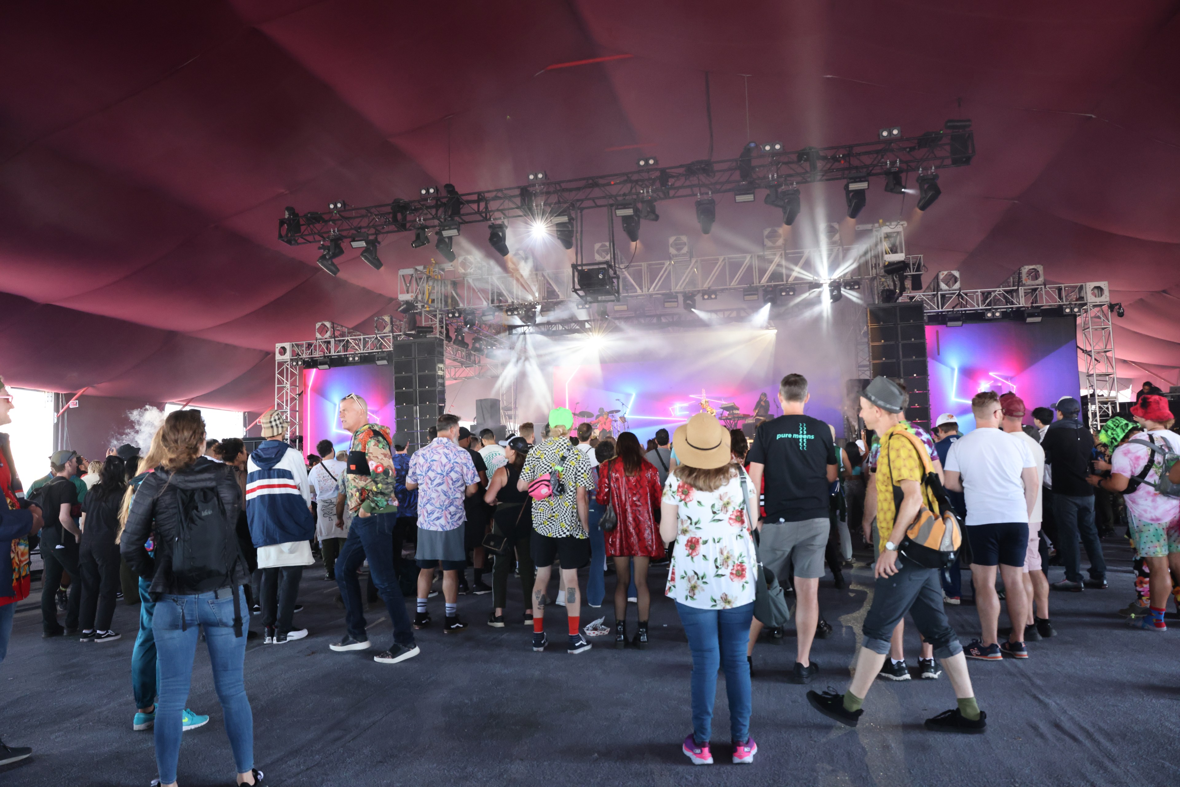 A crowd of people stands under a red tent, watching a brightly lit stage with screens and performers, while various lights and smoke effects enhance the atmosphere.