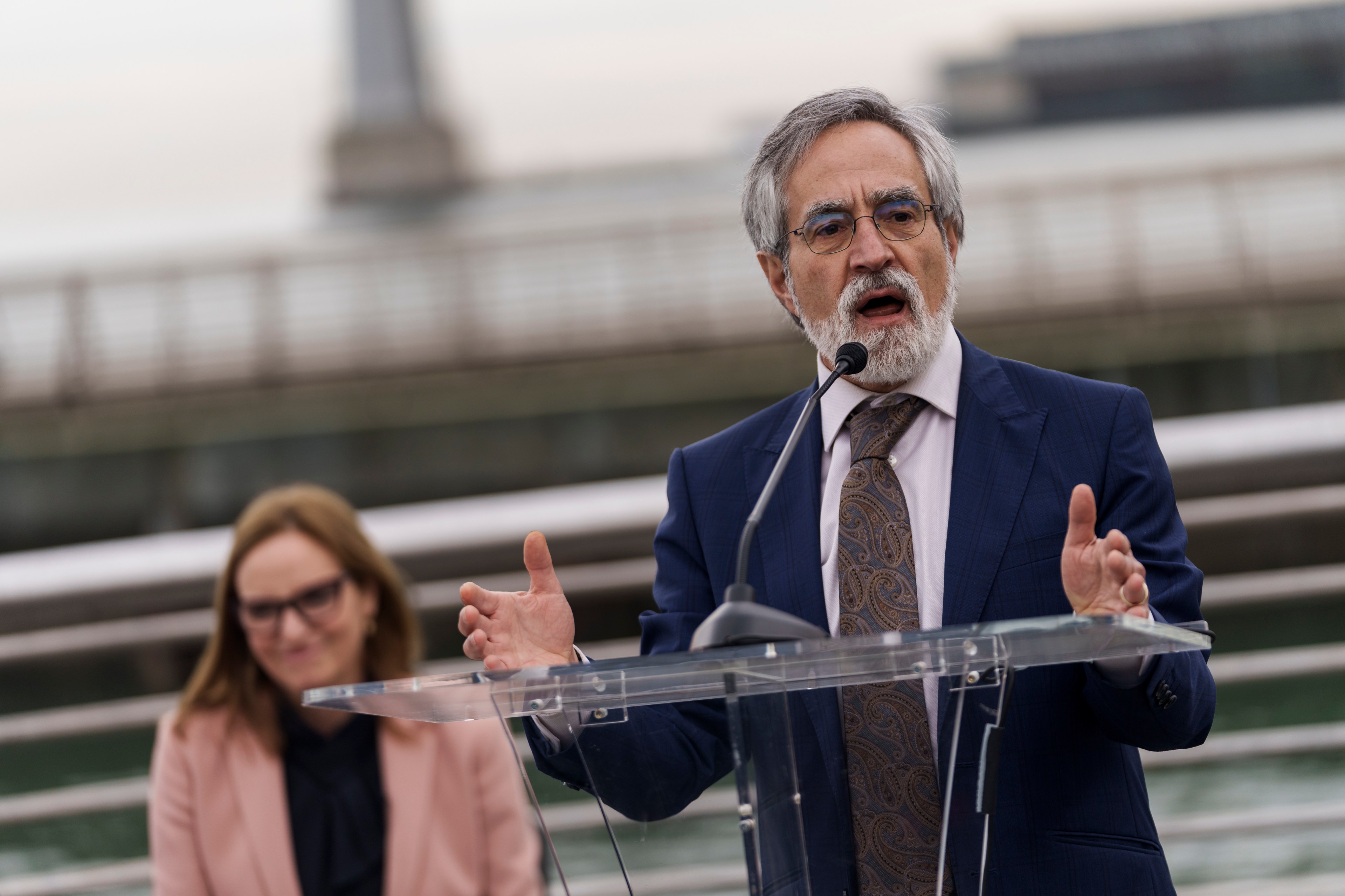 An older man with gray hair and a beard, wearing a suit, is speaking into a microphone at a podium. A woman in a pink blazer is seated in the background.