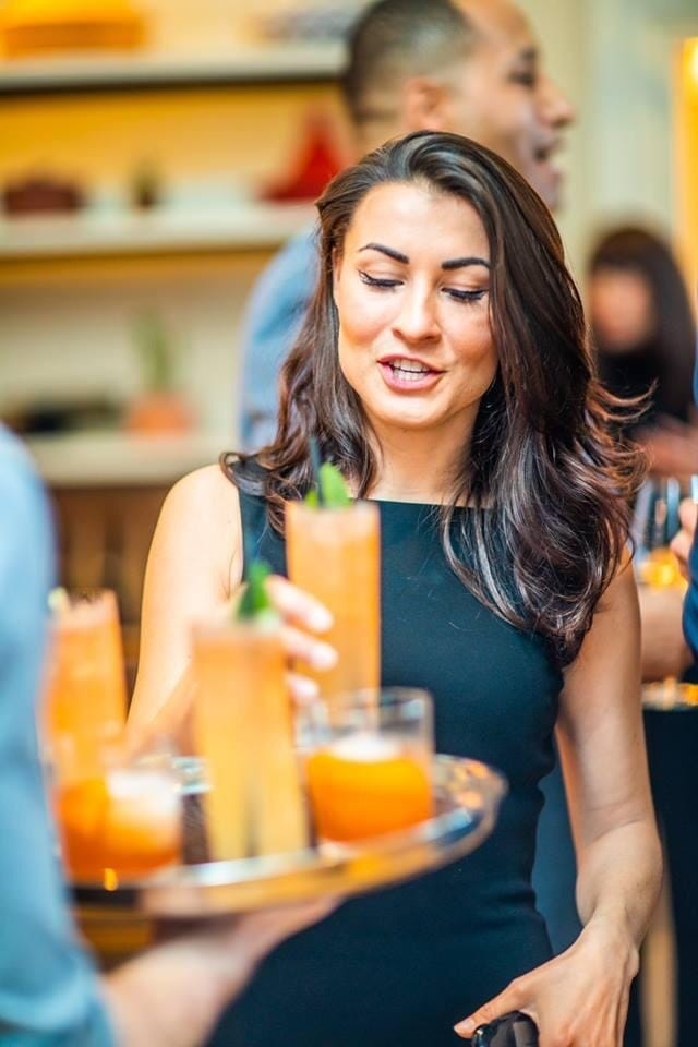 A woman in a black sleeveless dress is smiling and reaching for a tall cocktail glass from a tray held by another person, with blurred background of people and decor.