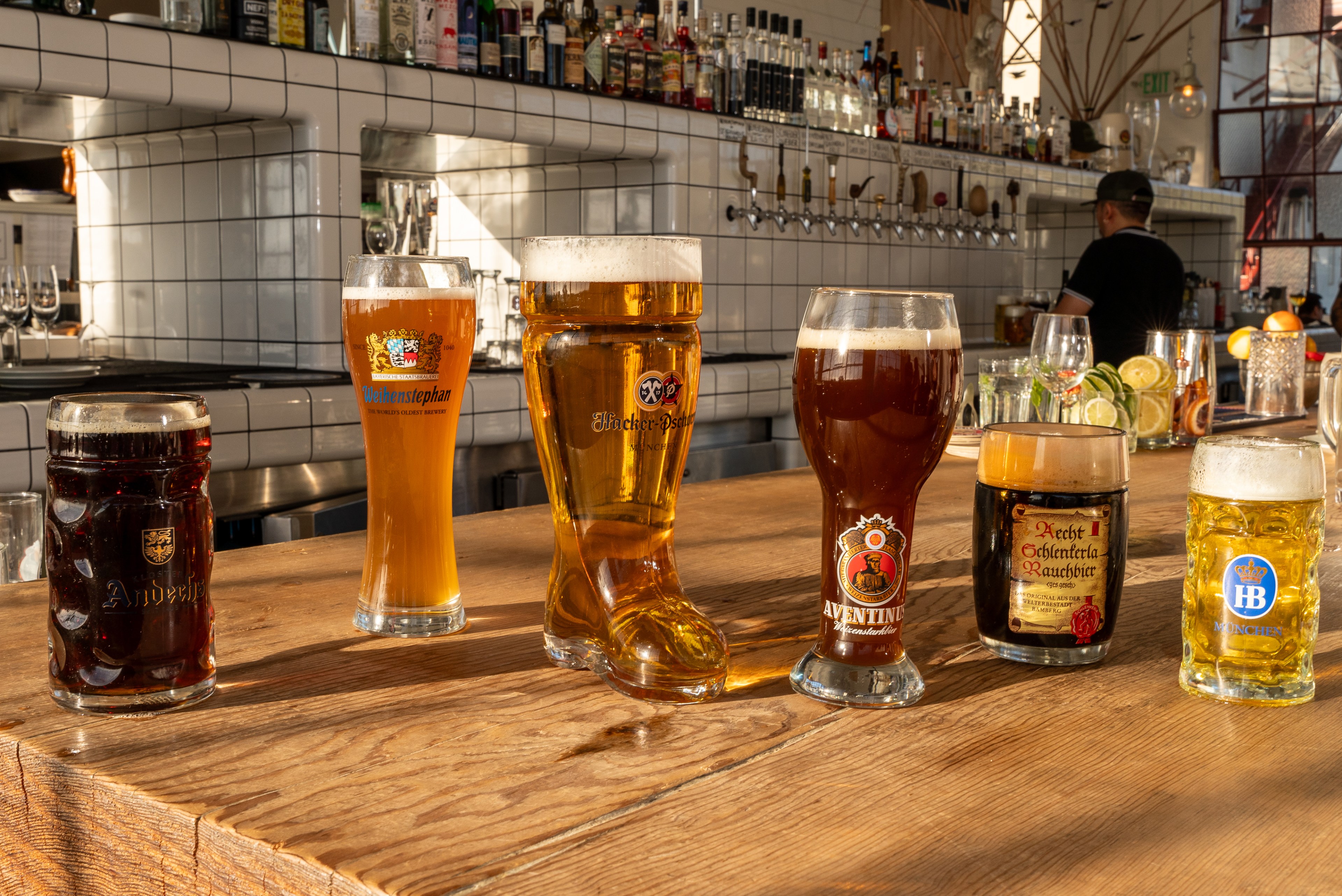 Six glasses of beer in varying styles and colors sit on a bar counter, with a well-stocked bar in the background and a person working behind it.