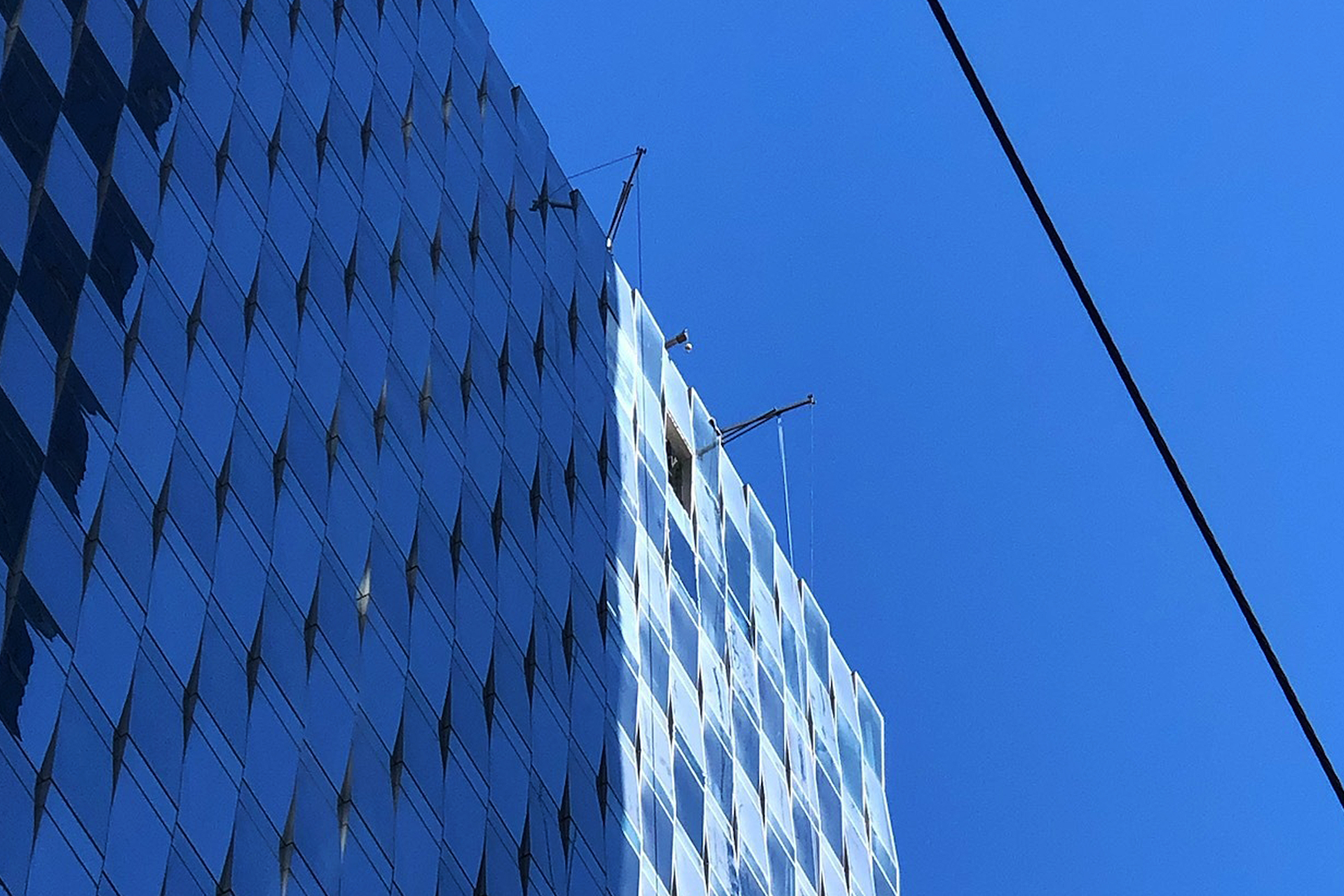 A glass-covered high-rise building reflects the blue sky, while two window cleaning platforms are visible near the top, with a cable diagonally across the scene.