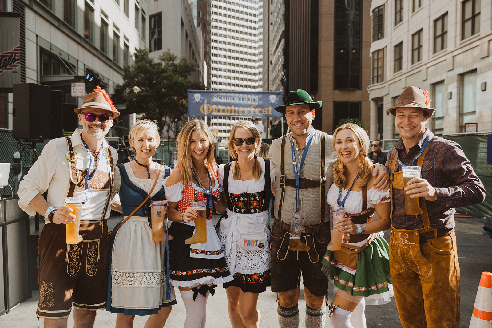 A group of seven people dressed in traditional Bavarian attire, holding drinks, stands on a city street, smiling for the camera. A sign behind them reads "Oktoberfest".