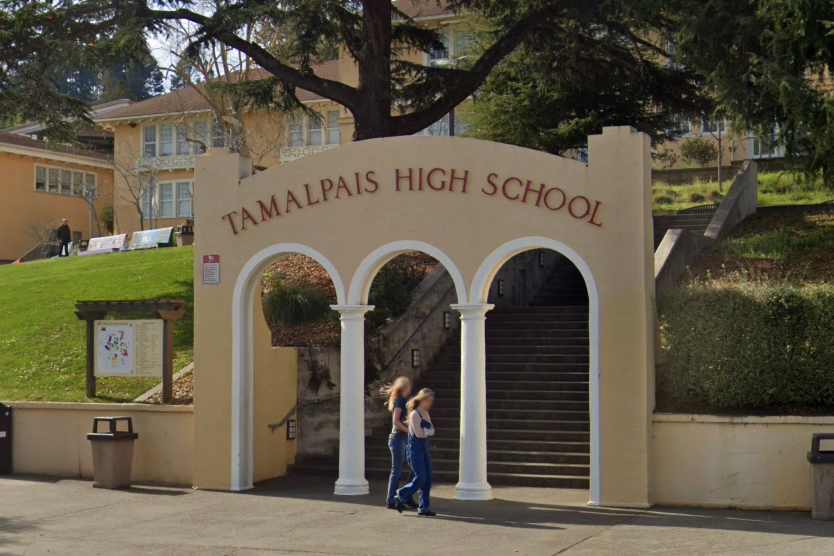 The image shows a beige archway with "Tamalpais High School" written on it, at the base of stairs leading up to a building surrounded by trees and greenery. Two people walk by.