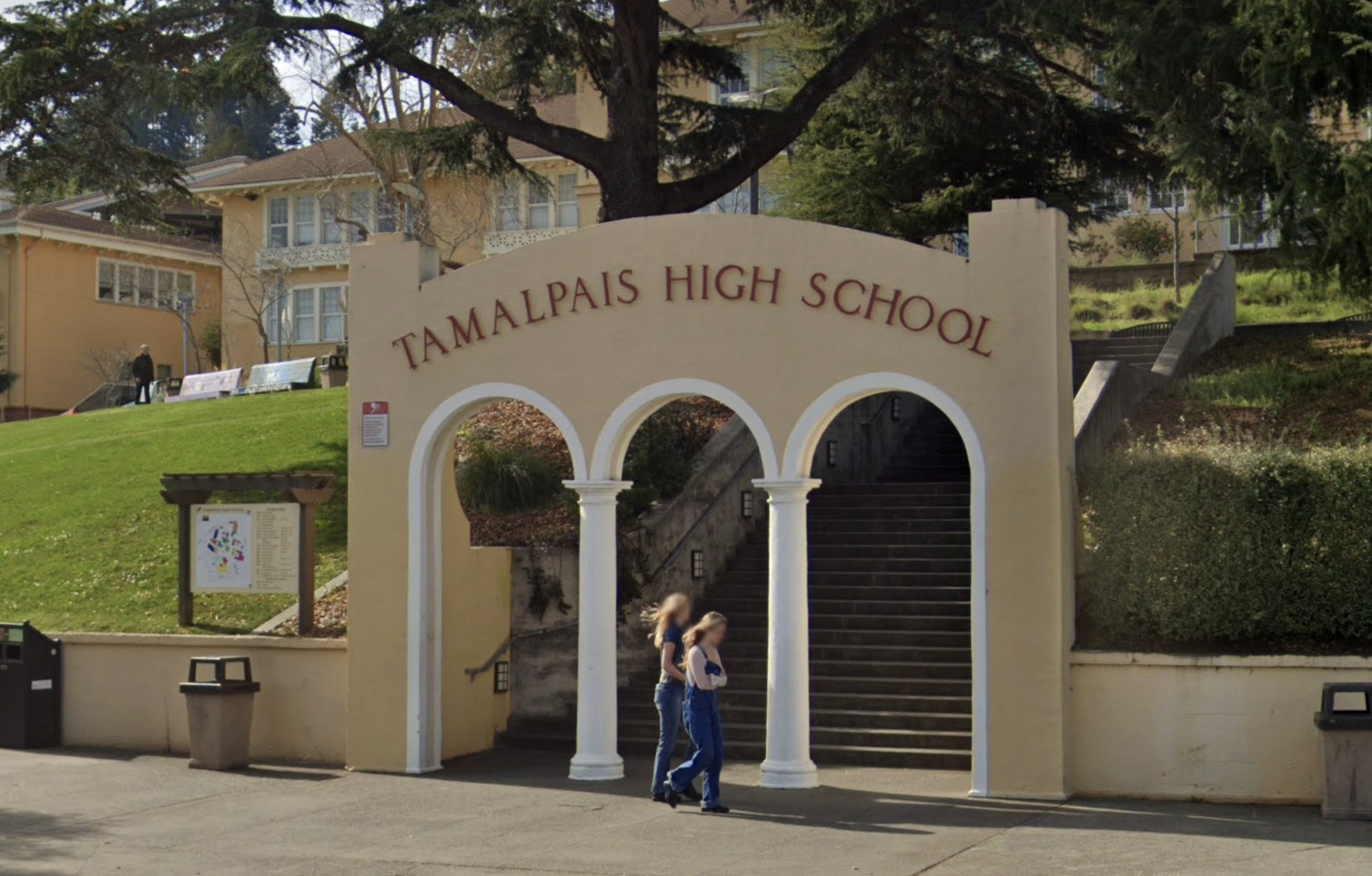 The image shows a beige archway with "Tamalpais High School" written on it, at the base of stairs leading up to a building surrounded by trees and greenery. Two people walk by.