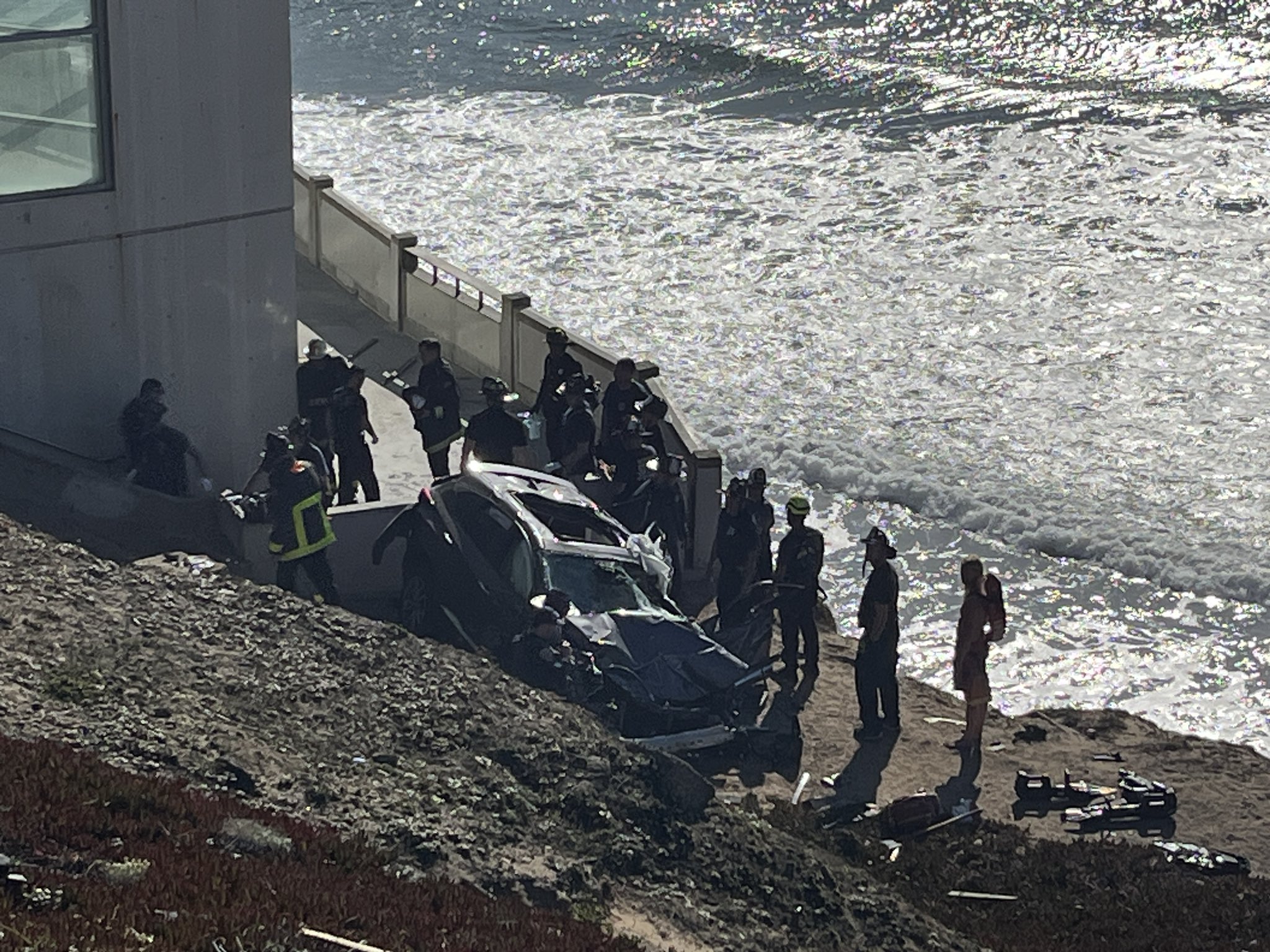 The image shows a car crash scene by the seaside, with emergency personnel gathered around a badly damaged vehicle at the base of a concrete structure.