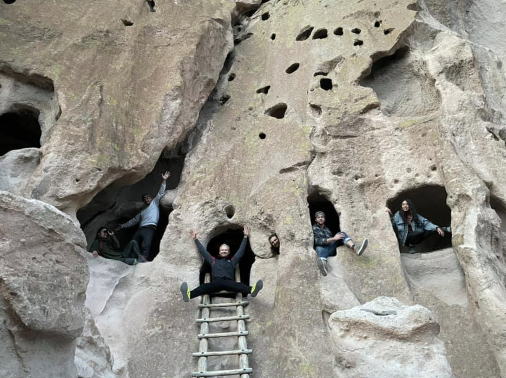 People are playfully sitting in small, cave-like openings in a large rock wall. They are smiling and posing, using a wooden ladder and surrounding rock formations.