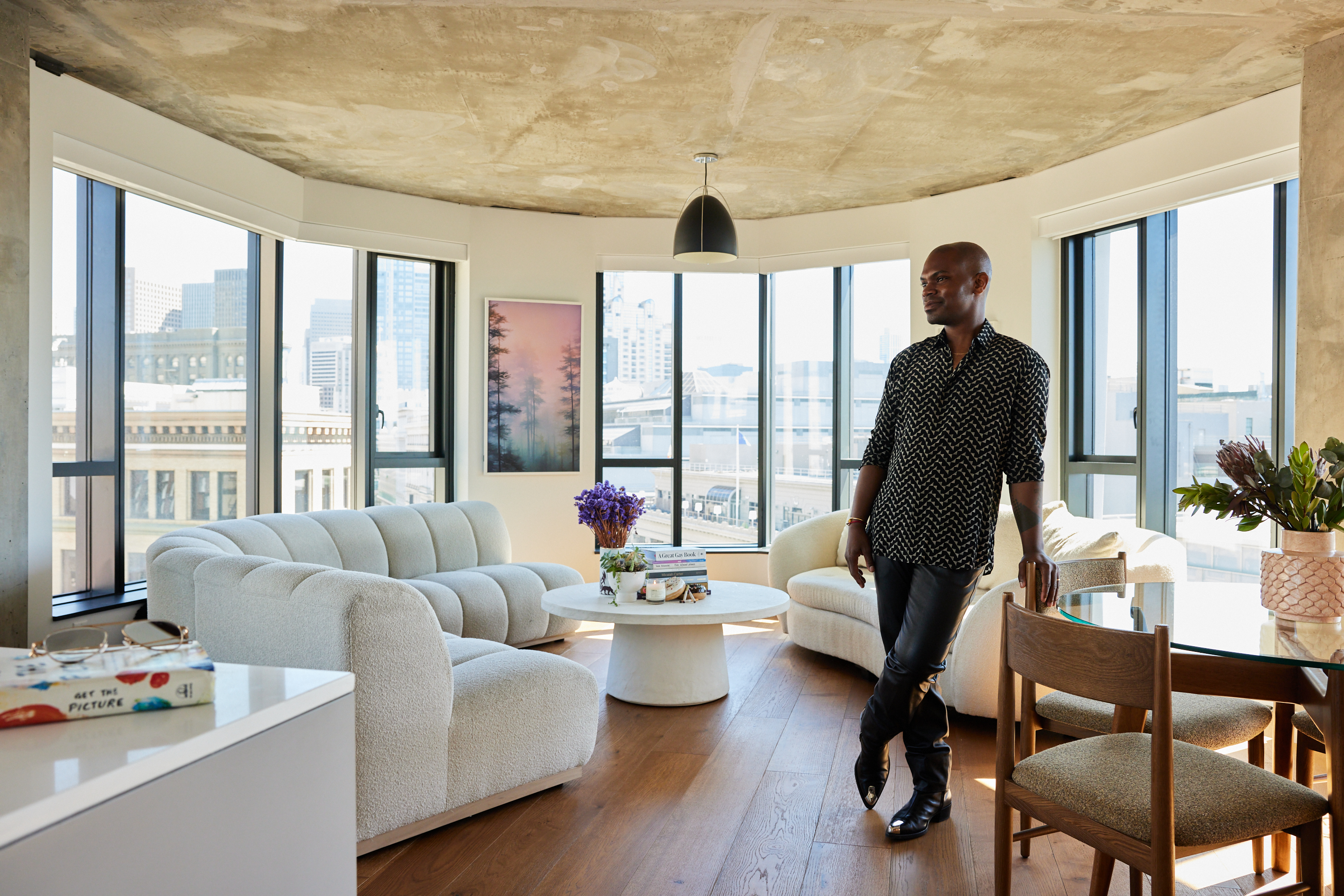 A person stands in a stylish, sunlit living room with large windows, white modern furniture, and wooden flooring. There's a cityscape in the background.