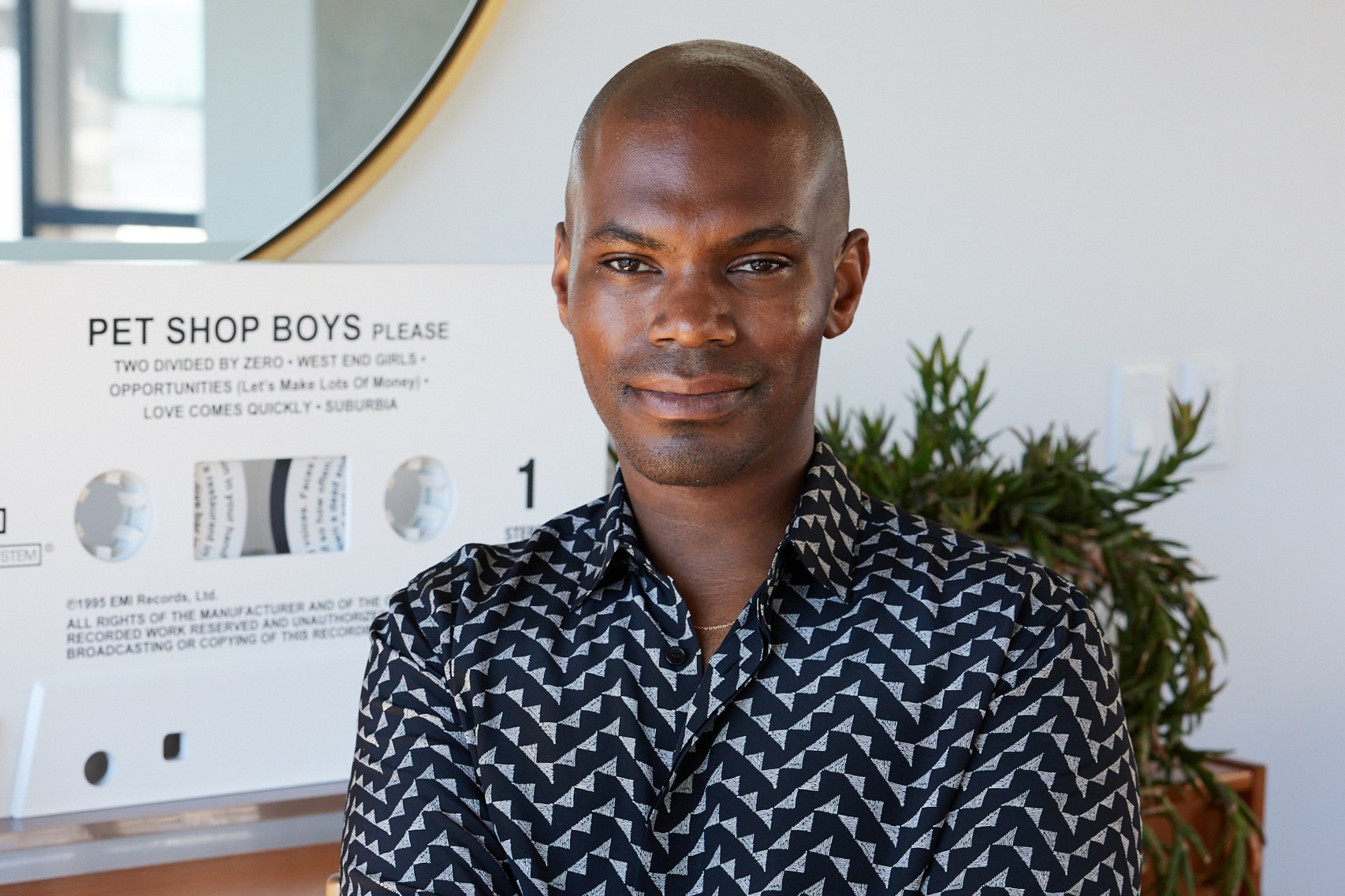 A person sits with an arm crossed, wearing a patterned shirt. Behind is a large circular mirror and a "Pet Shop Boys" sign. There’s also a plant in the background.