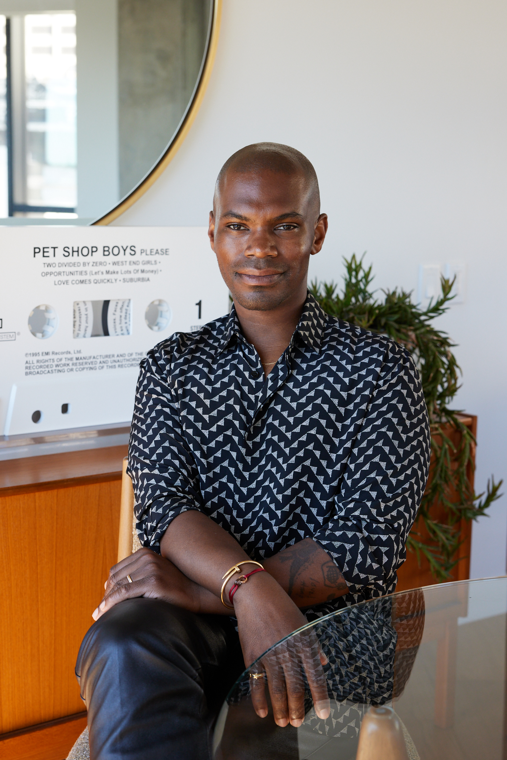 A person sits with an arm crossed, wearing a patterned shirt. Behind is a large circular mirror and a "Pet Shop Boys" sign. There’s also a plant in the background.