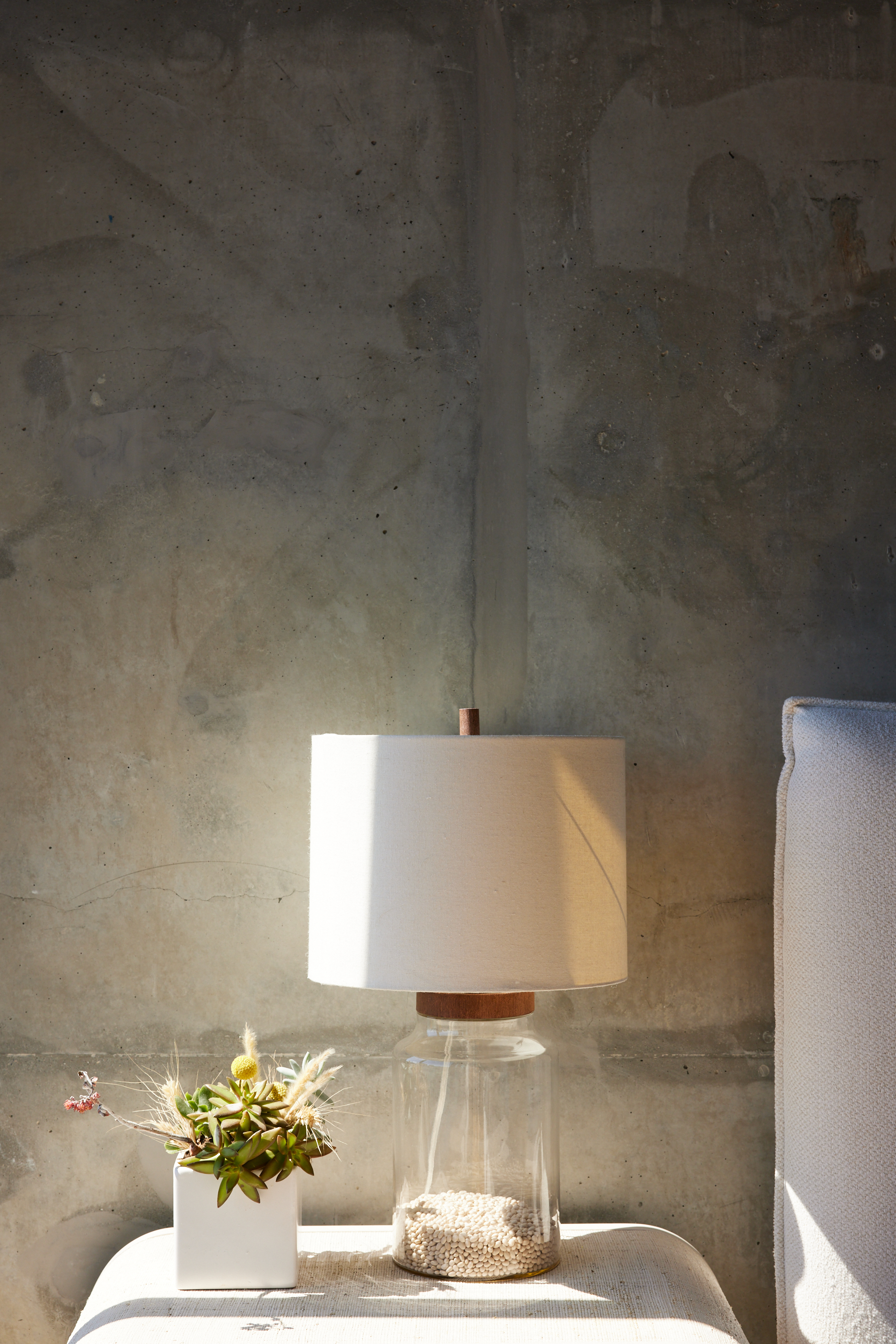 The image shows a minimalist setting with a glass lamp containing white pebbles and a white shade. Next to it is a small potted succulent, all against a grey concrete wall.