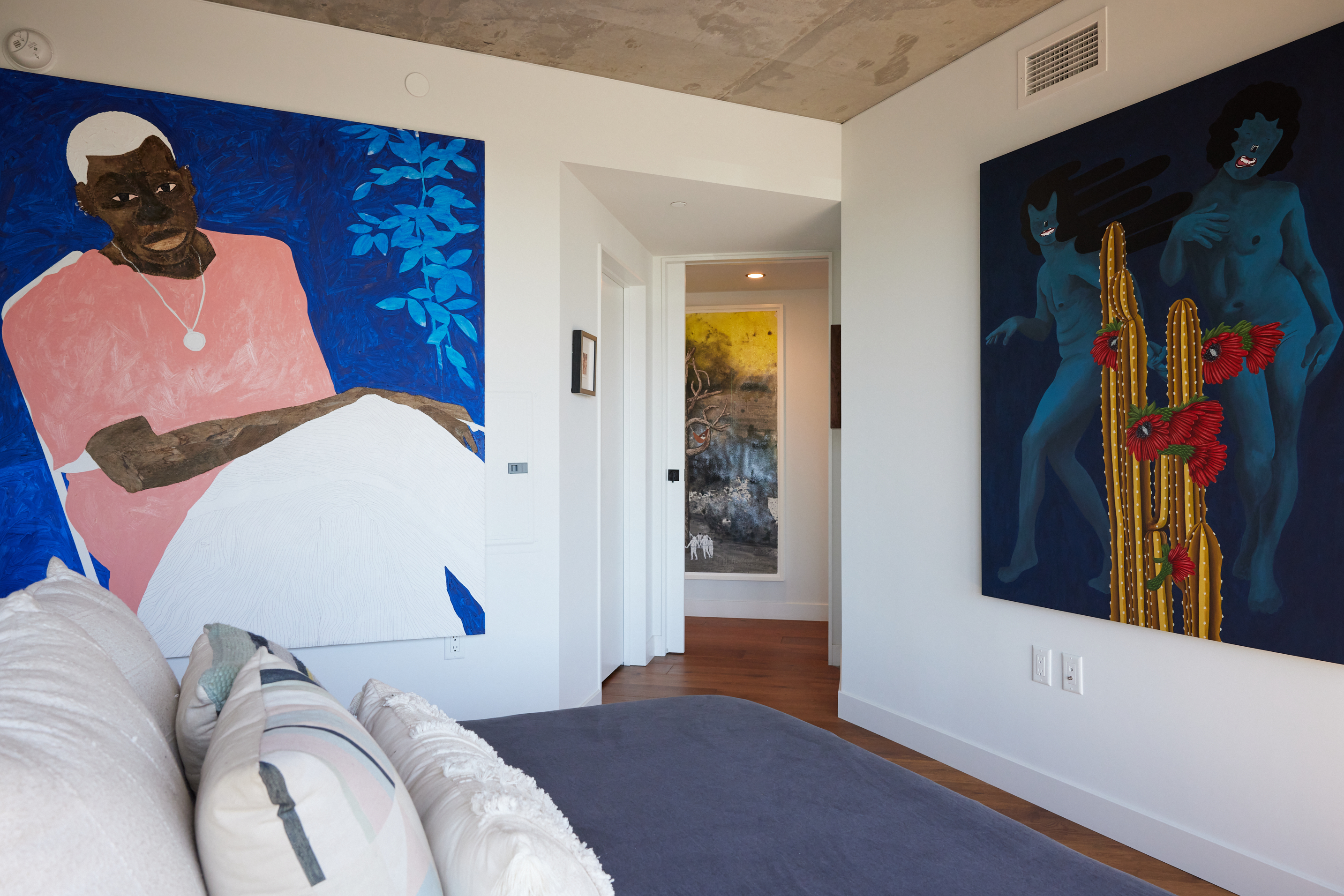 The image shows a modern room with two large, vibrant paintings on the walls, a plush gray bed with white and patterned pillows, and a hallway leading to more artwork.