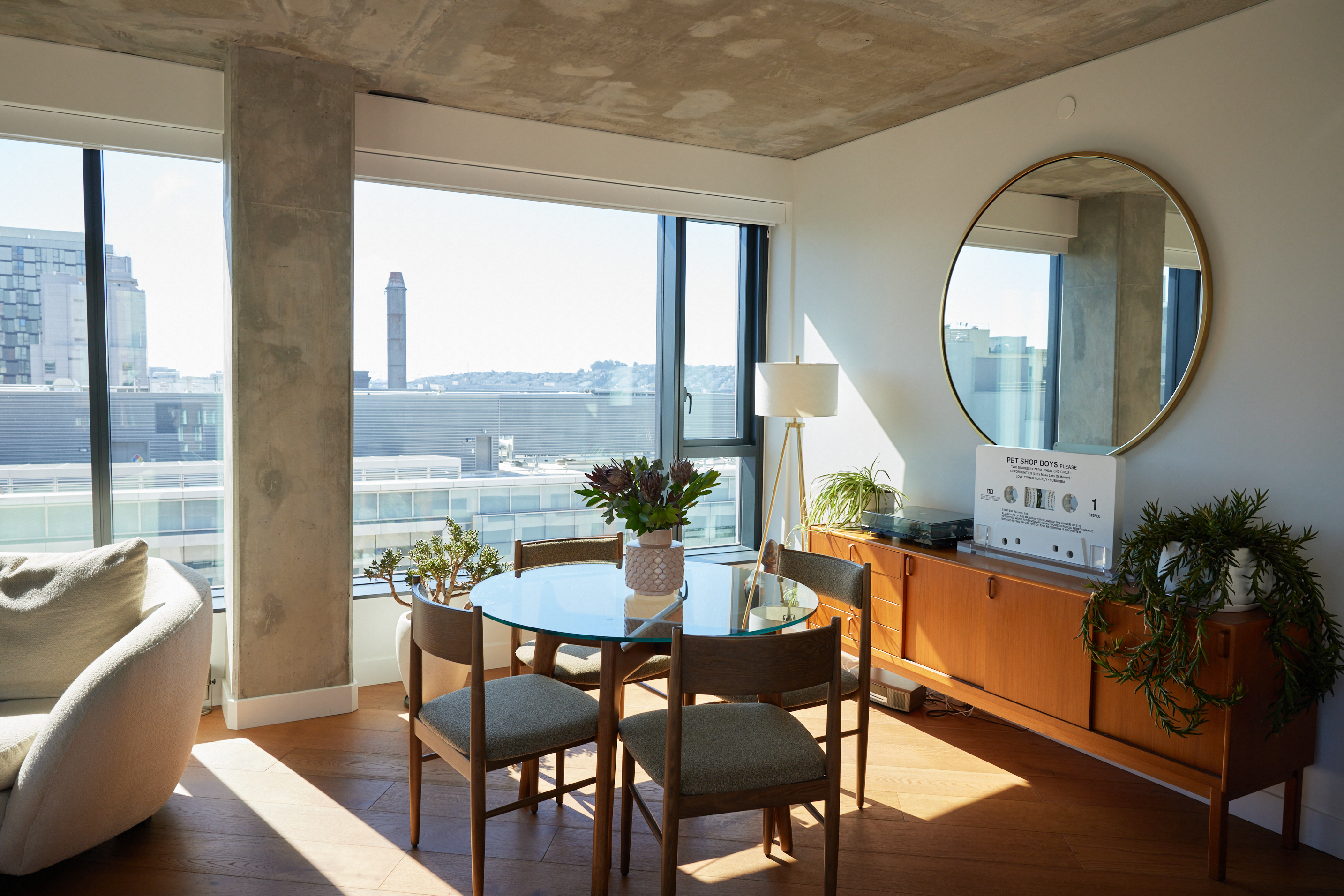 A cozy modern apartment with large windows, a round mirror, a glass dining table with four chairs, plants, and a sideboard with a lamp and a stereo.