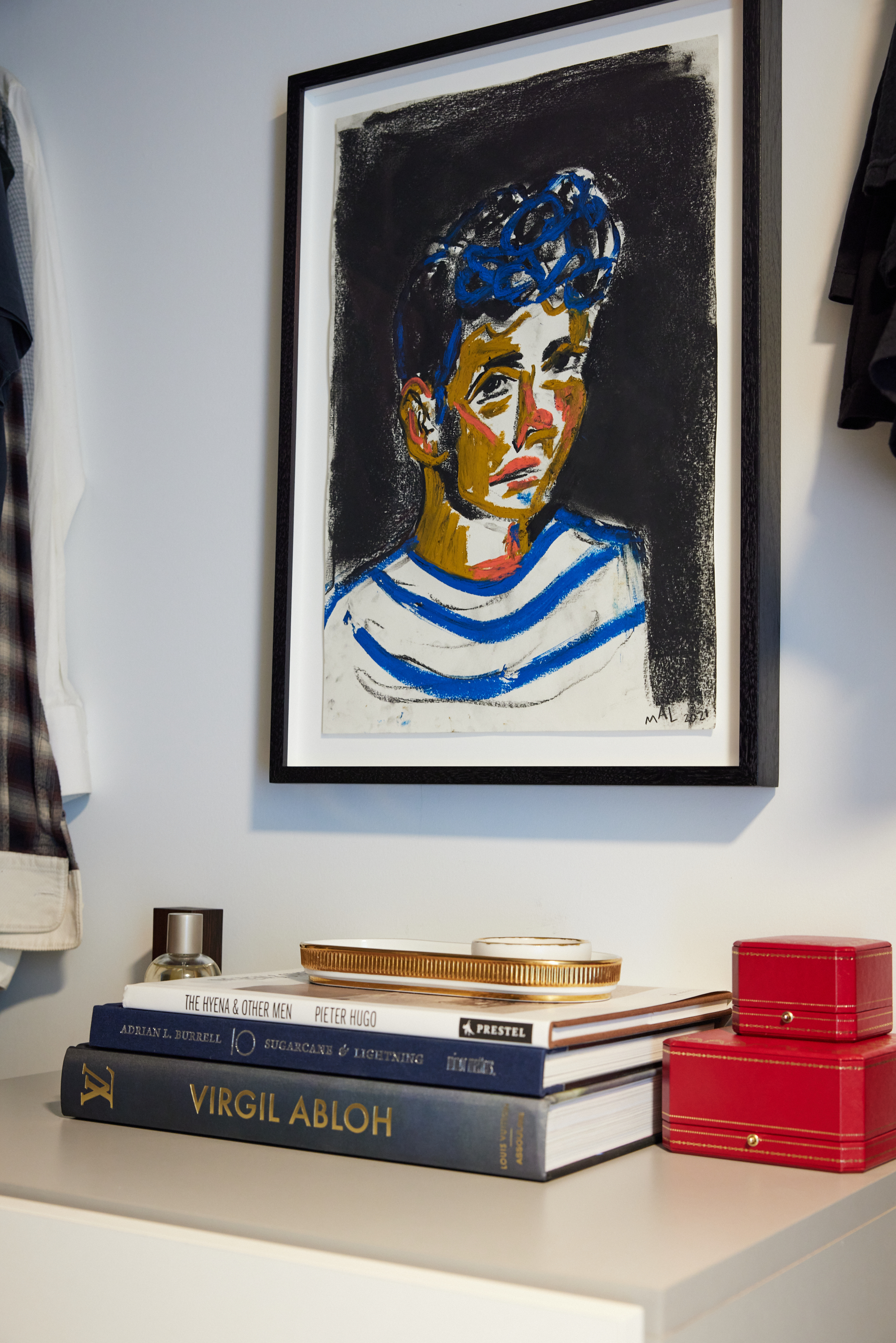 The image shows a framed abstract painting of a person with curly hair in a striped shirt, hung above a stack of books and red boxes on a dresser.