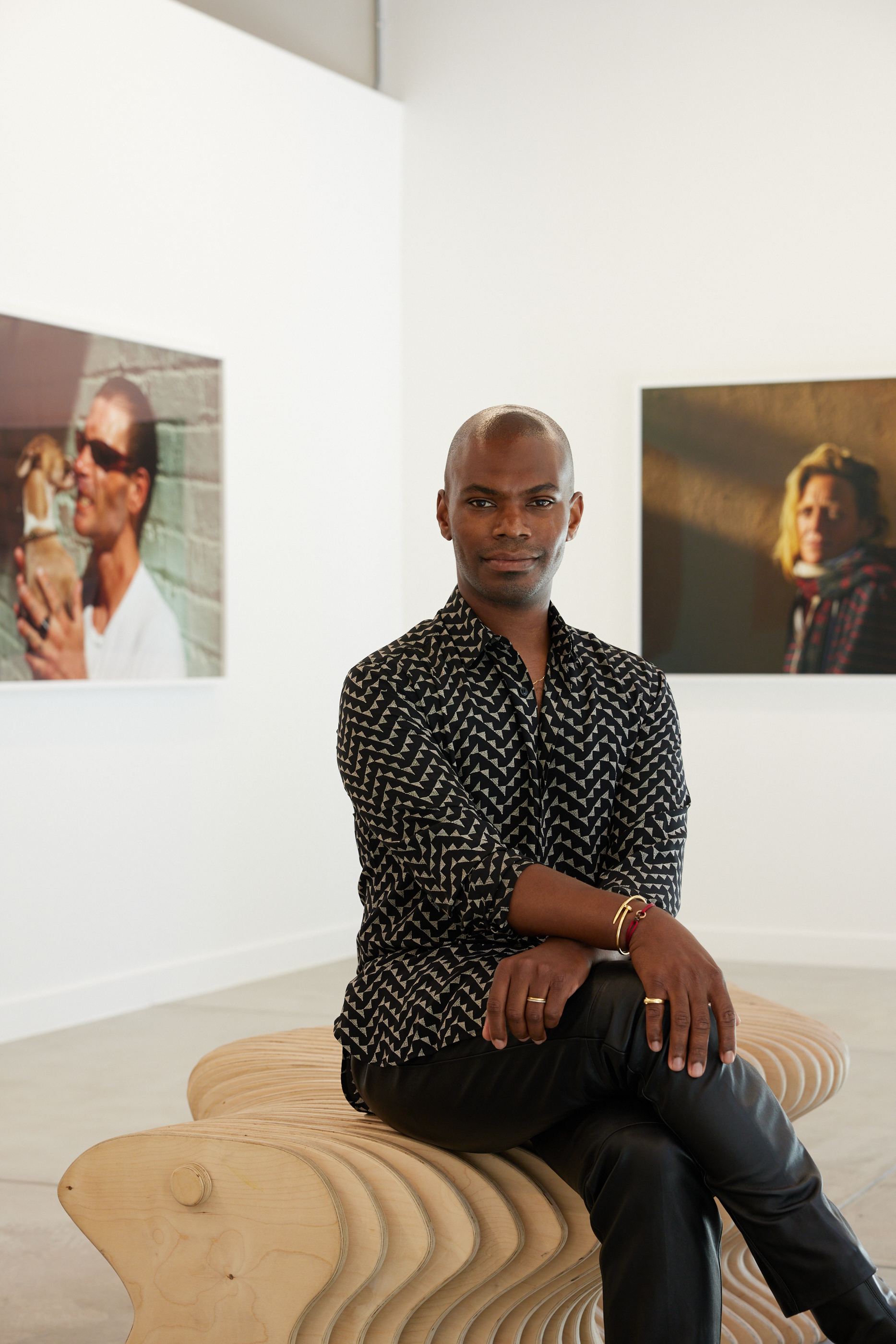 A person with a patterned shirt and bracelet sits on a wooden bench in a gallery with two framed photos on the wall behind them.