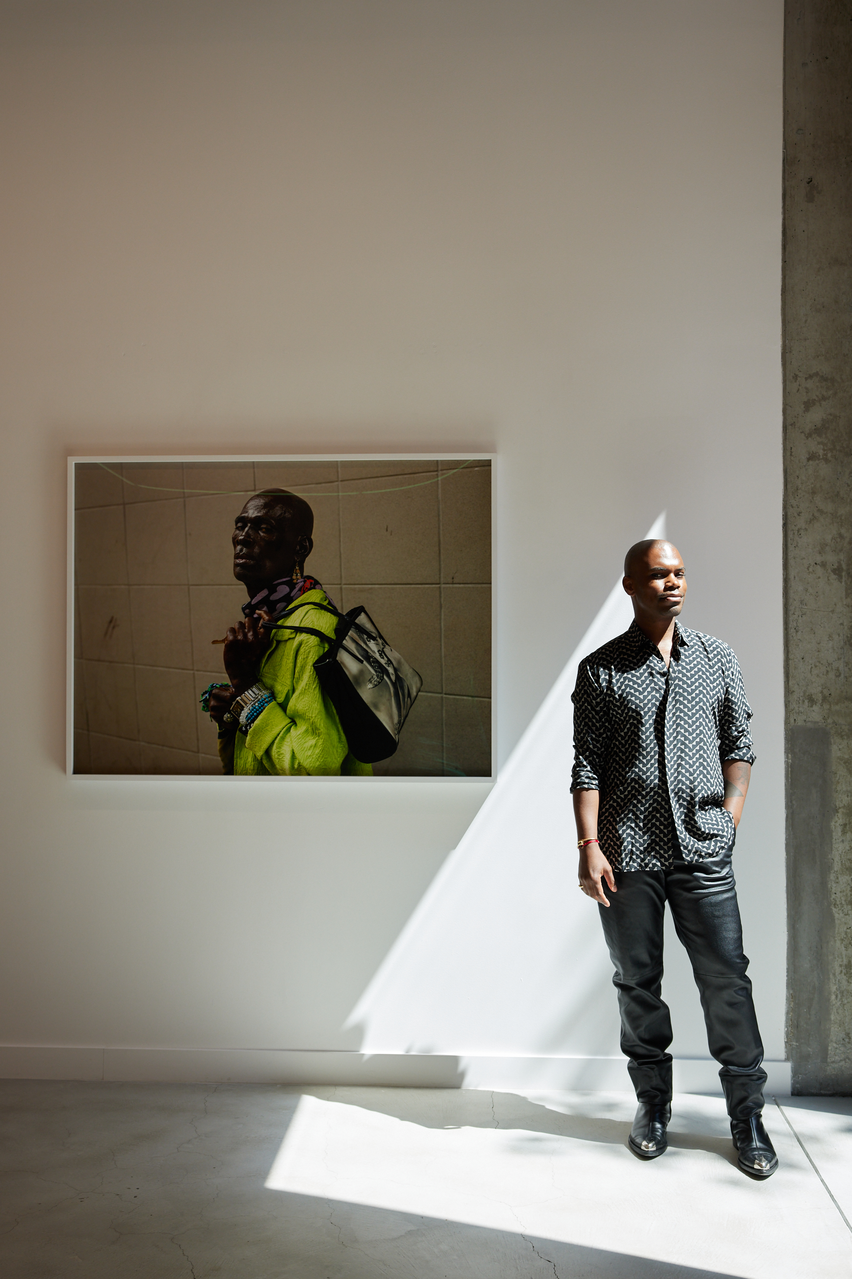 A man in a patterned shirt and dark pants stands beside a framed photo of another man in a green jacket with a bag. Sunlight creates a diagonal shadow on the wall.