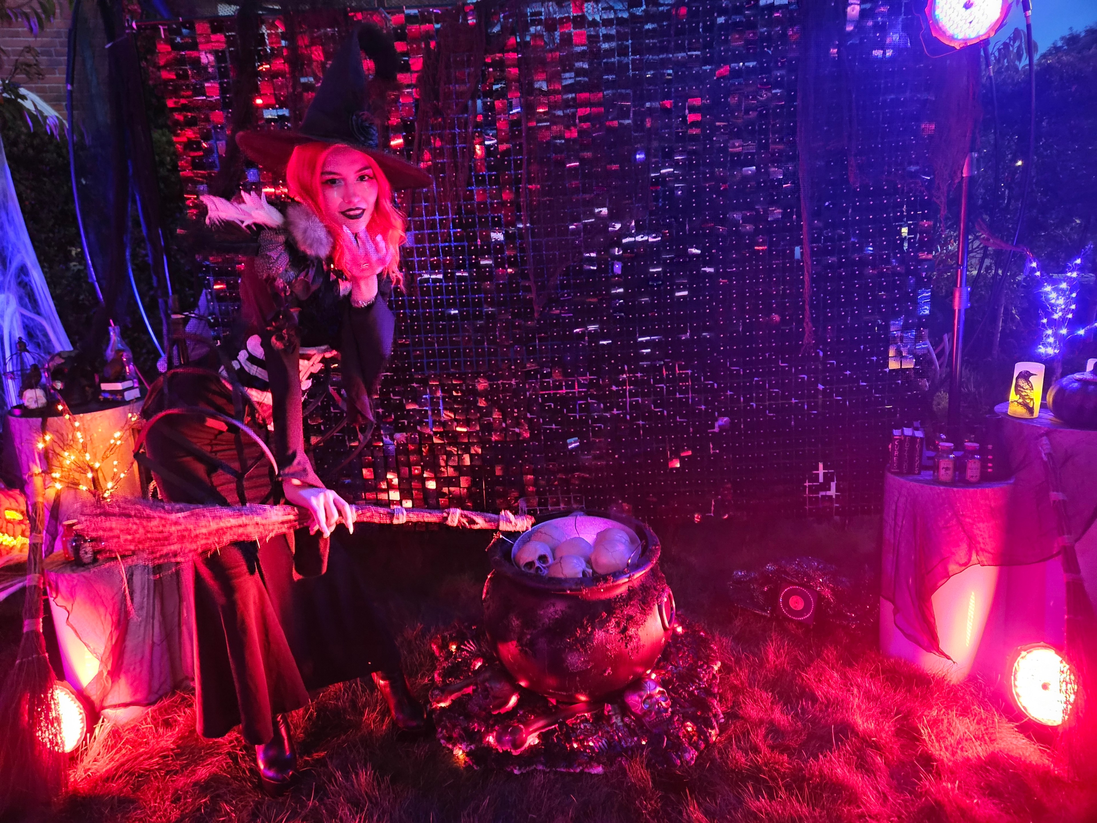 A woman dressed as a witch is posing with a broom in a Halloween scene. She stands next to a cauldron with skulls, surrounded by spooky decorations and eerie lighting.