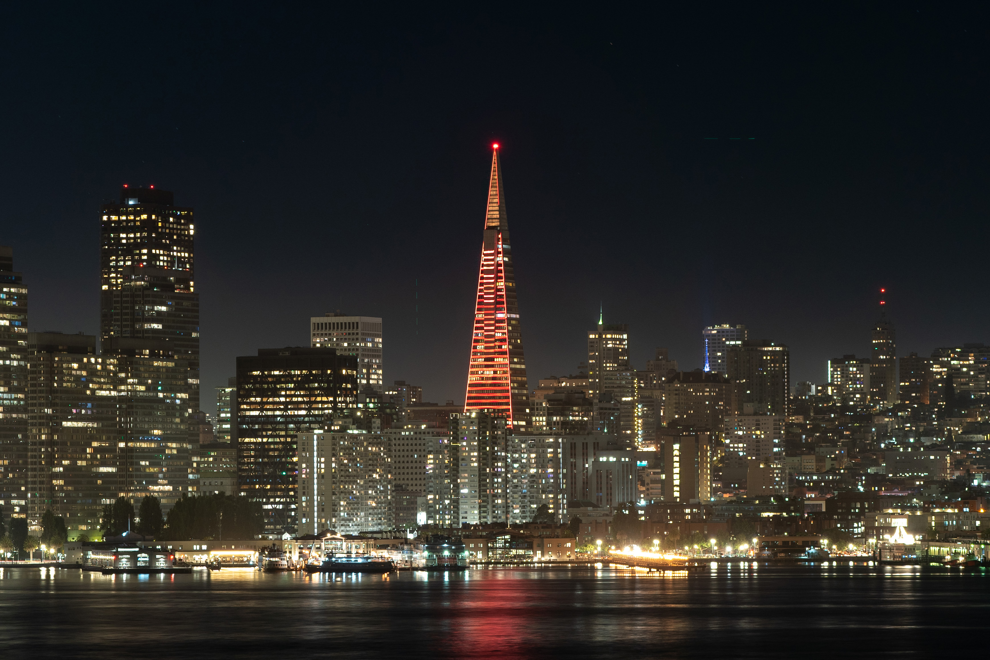 The image shows a city skyline at night with a tall, pyramid-shaped building illuminated in red at the center, surrounded by other lit-up buildings, with water in the foreground.