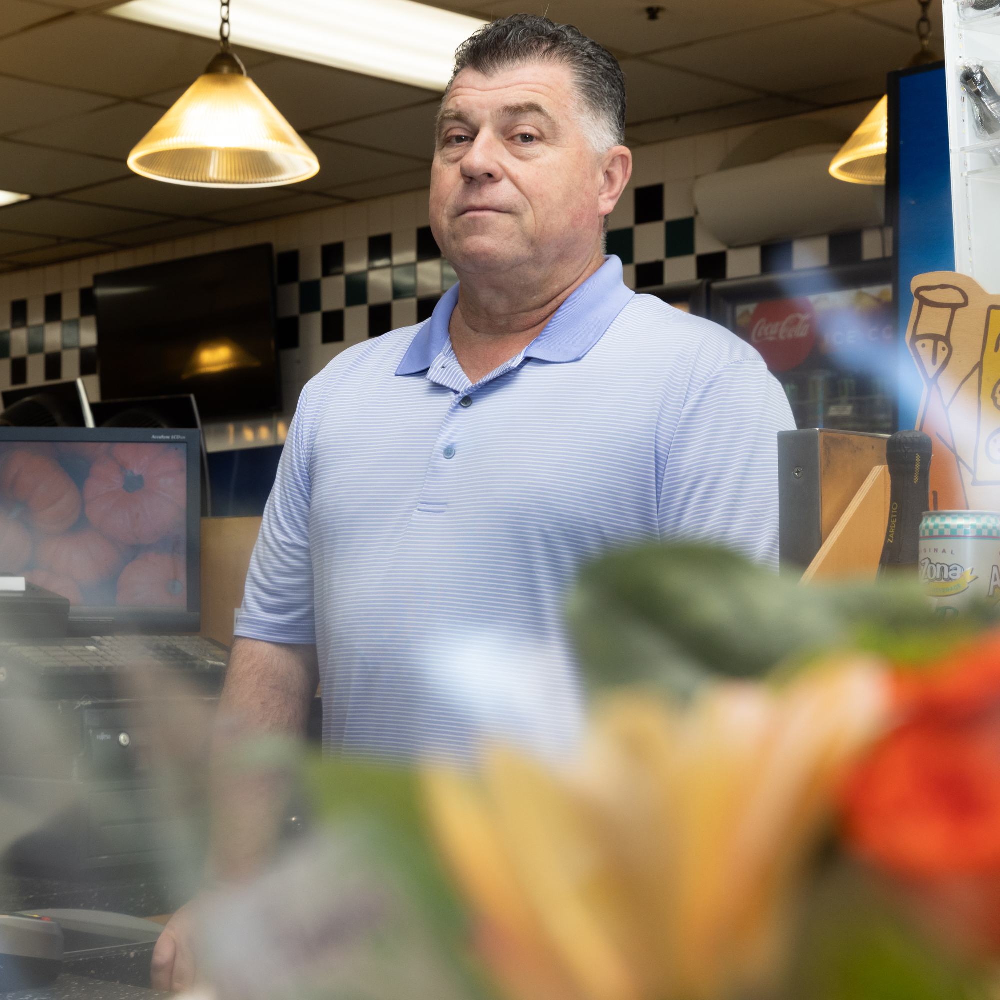 A man in a light blue polo shirt stands behind a counter in a tiled store; a computer screen with pumpkins and blurred flowers are visible.