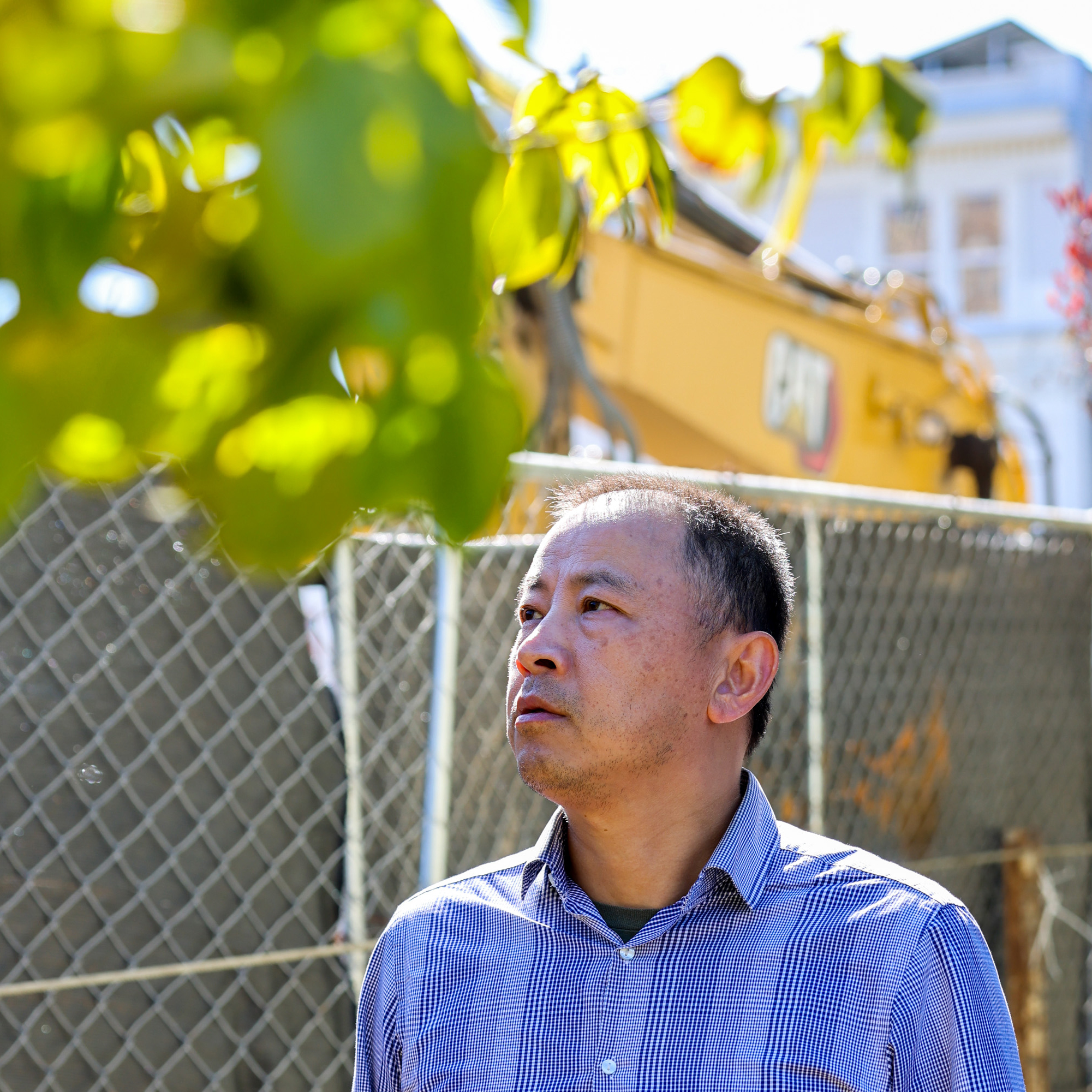 A man in a checkered shirt is standing in front of a fence, looking up. A yellow construction vehicle and blurred leaves are visible in the background.