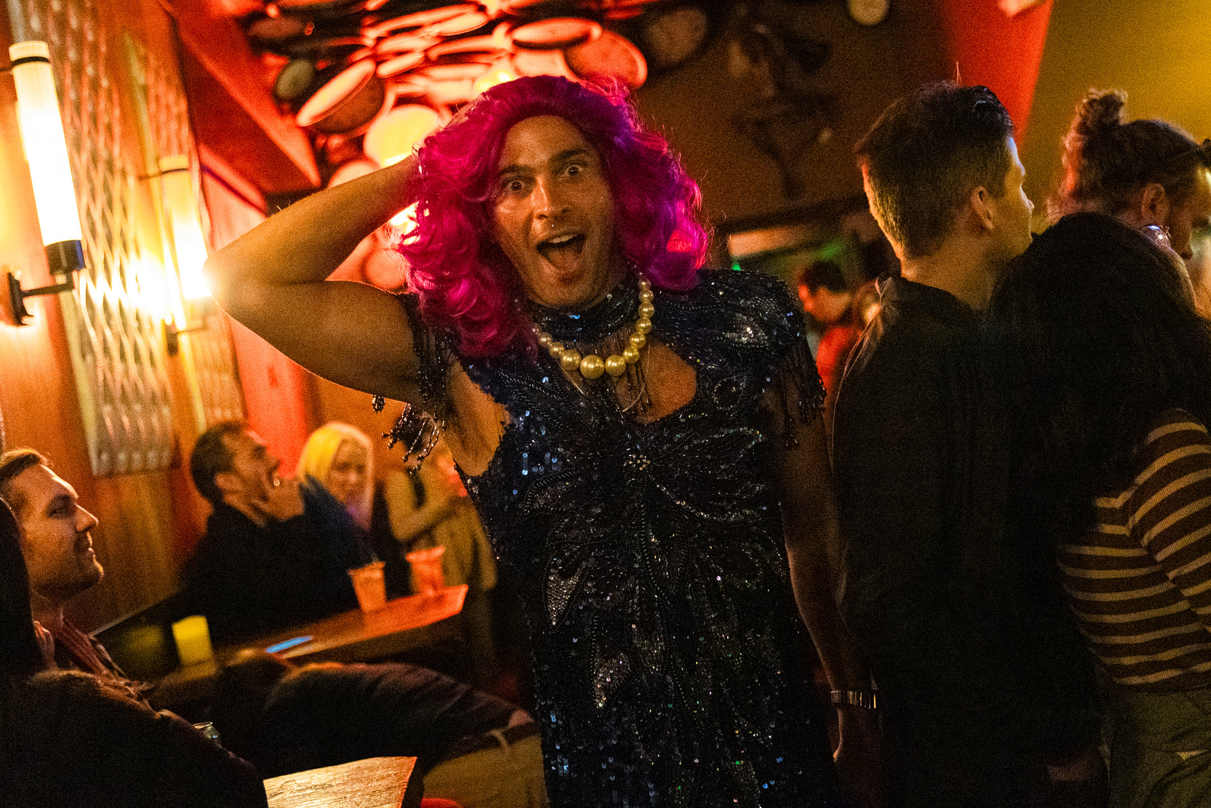 A person dressed in an extravagant outfit with a pink wig and a necklace gestures dramatically in a lively, crowded room with warm lighting and engaged patrons.