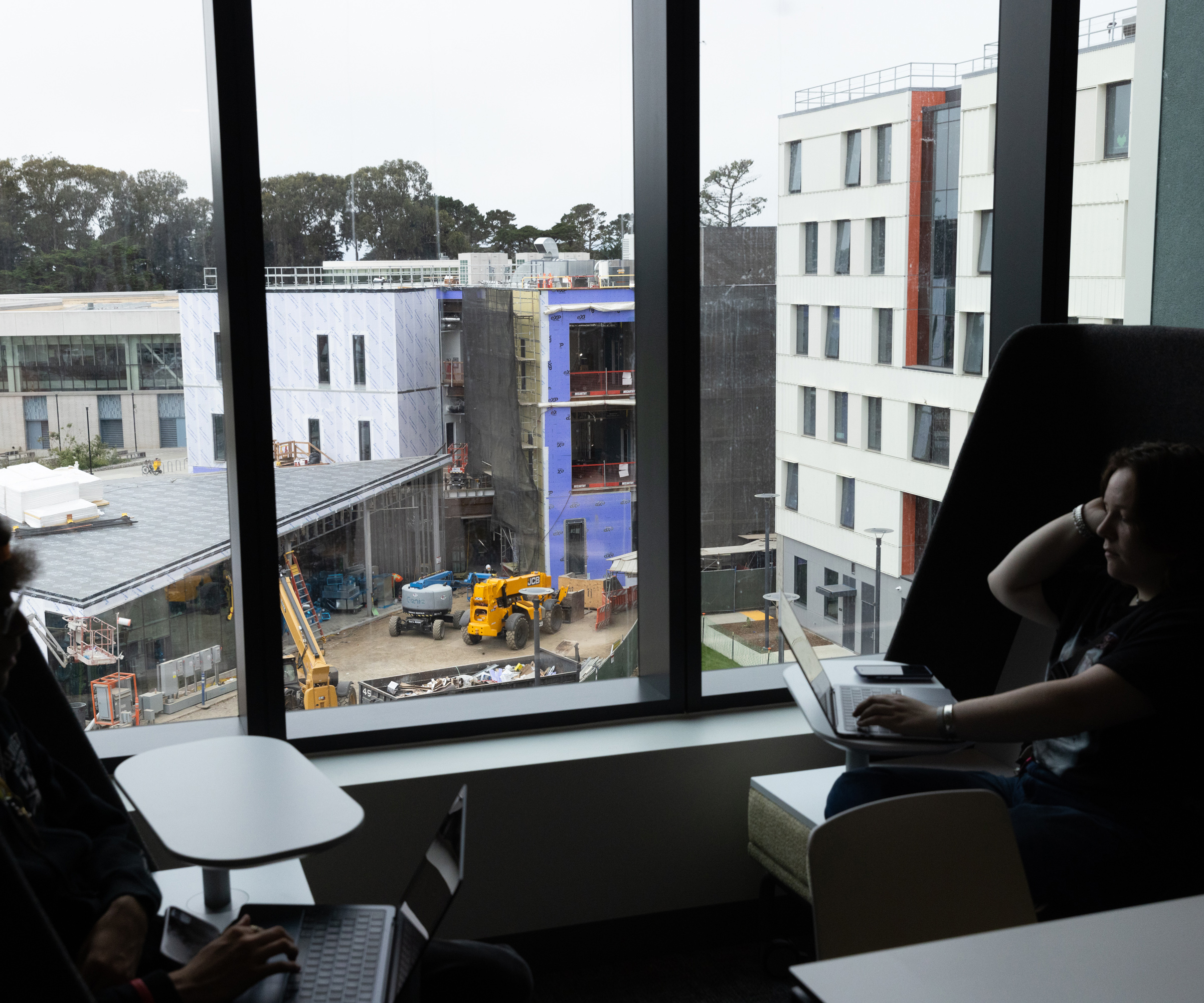 Two people are working on laptops near large windows overlooking a construction site with several vehicles and partially built structures.