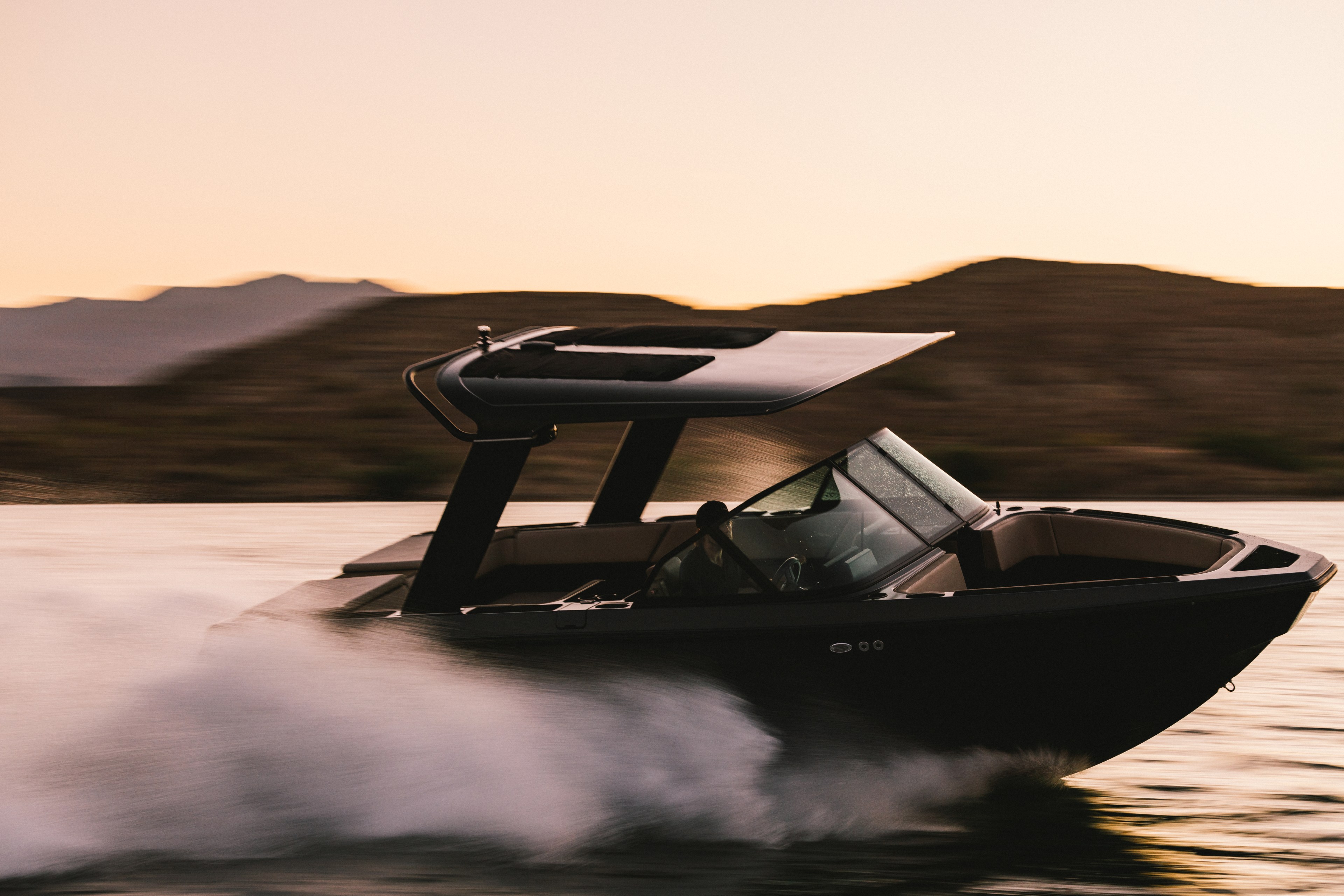 A sleek, modern speedboat glides swiftly over the water at sunset, creating a spray behind it, with rugged hills in the background.