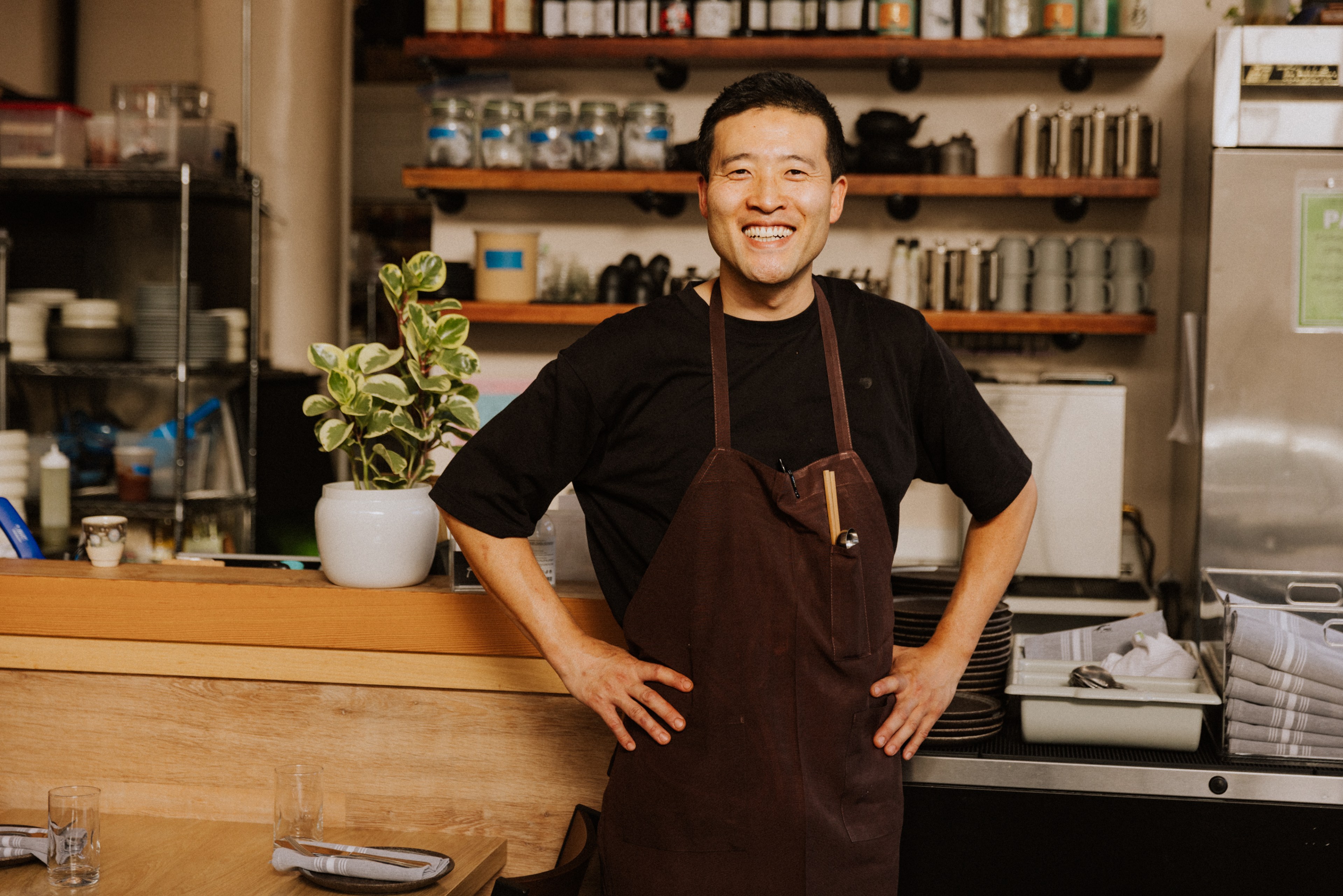 A smiling person in a dark apron stands in a warmly lit kitchen, hands on hips. Behind them are shelves with jars and kitchen items, and a potted plant nearby.