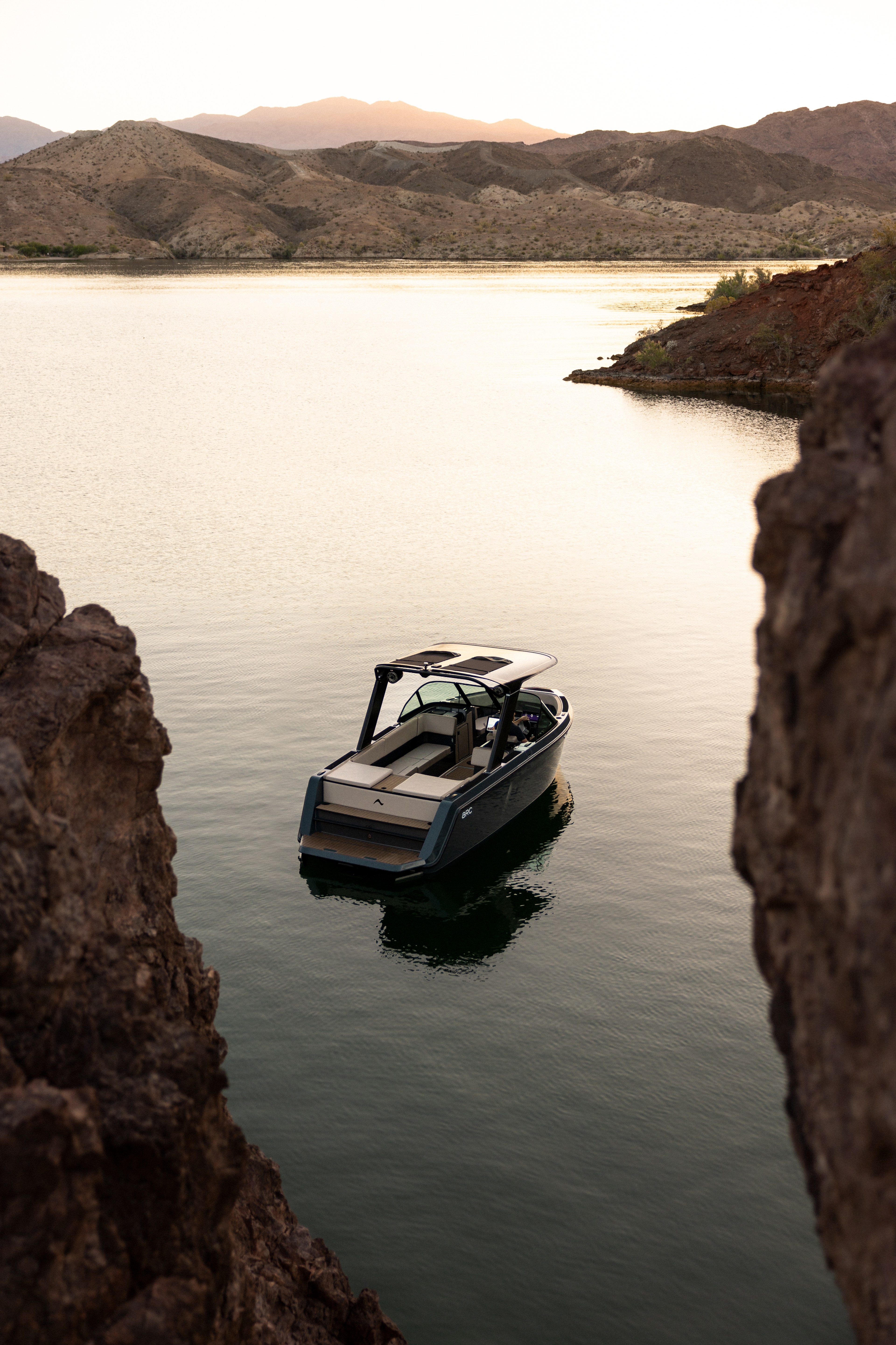 A sleek, modern boat floats on calm water, framed by rocky outcrops, set against a backdrop of rolling hills at sunset.