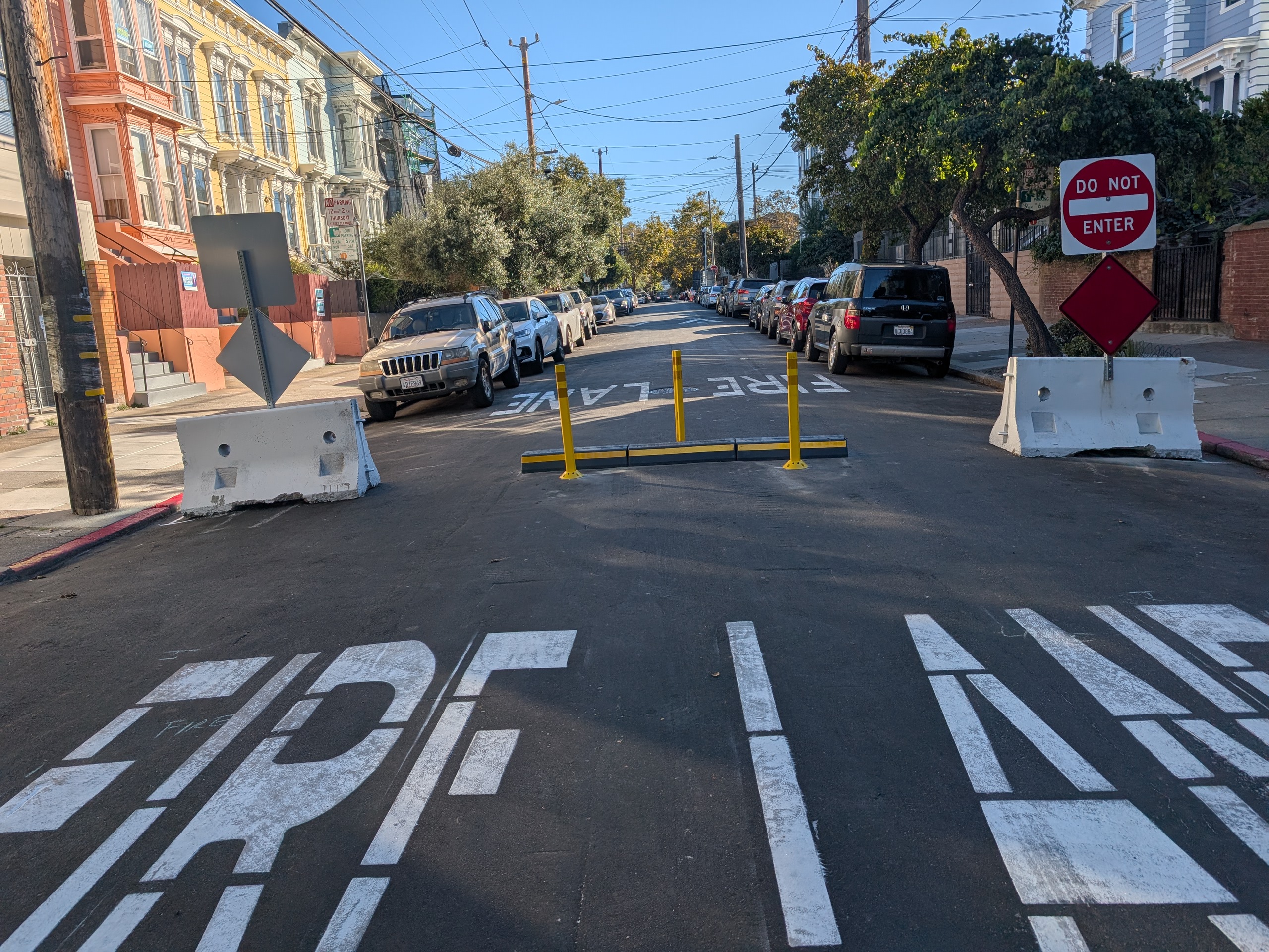 The image shows a street with &quot;FIRE LANE&quot; painted on it, blocked by barriers and a &quot;DO NOT ENTER&quot; sign. Parked cars line both sides of the street.