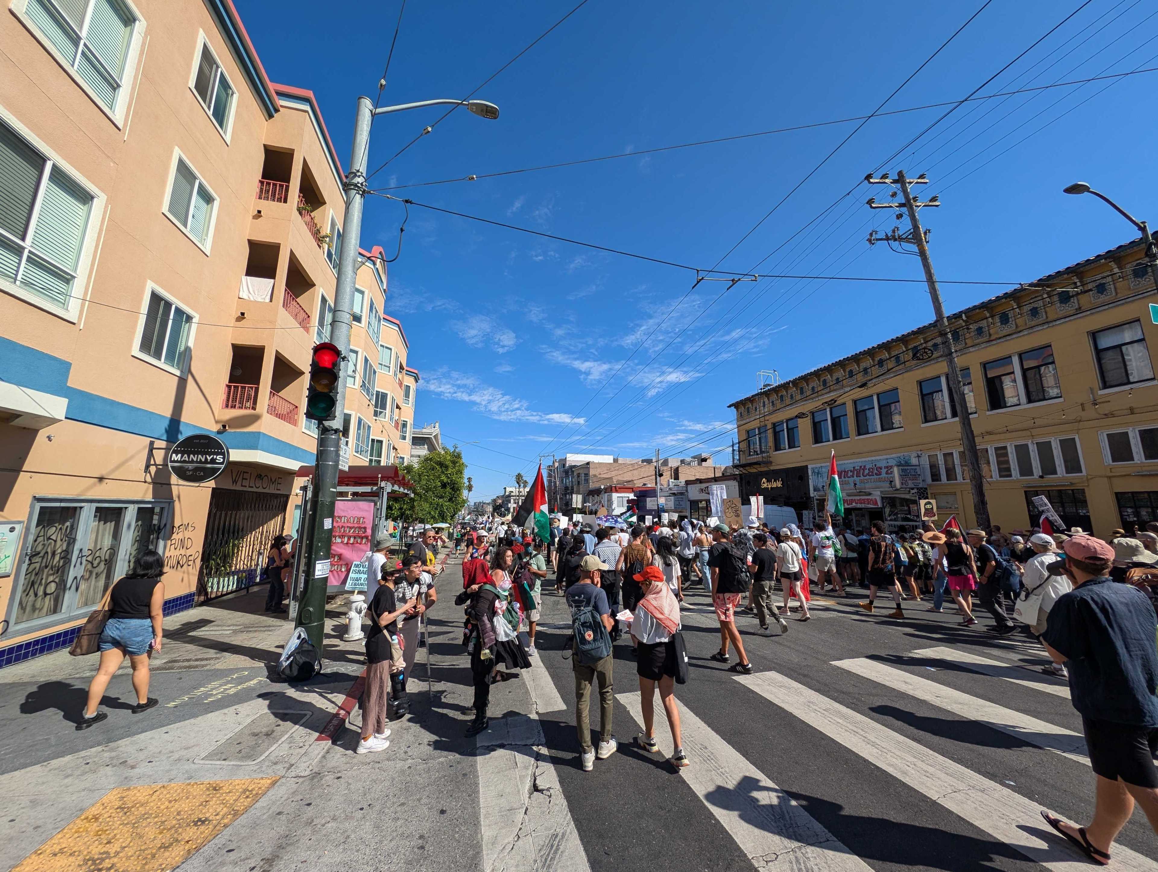 A crowd of people is marching down a city street under a clear blue sky, holding signs and flags. Buildings line both sides of the street, with a traffic light visible.