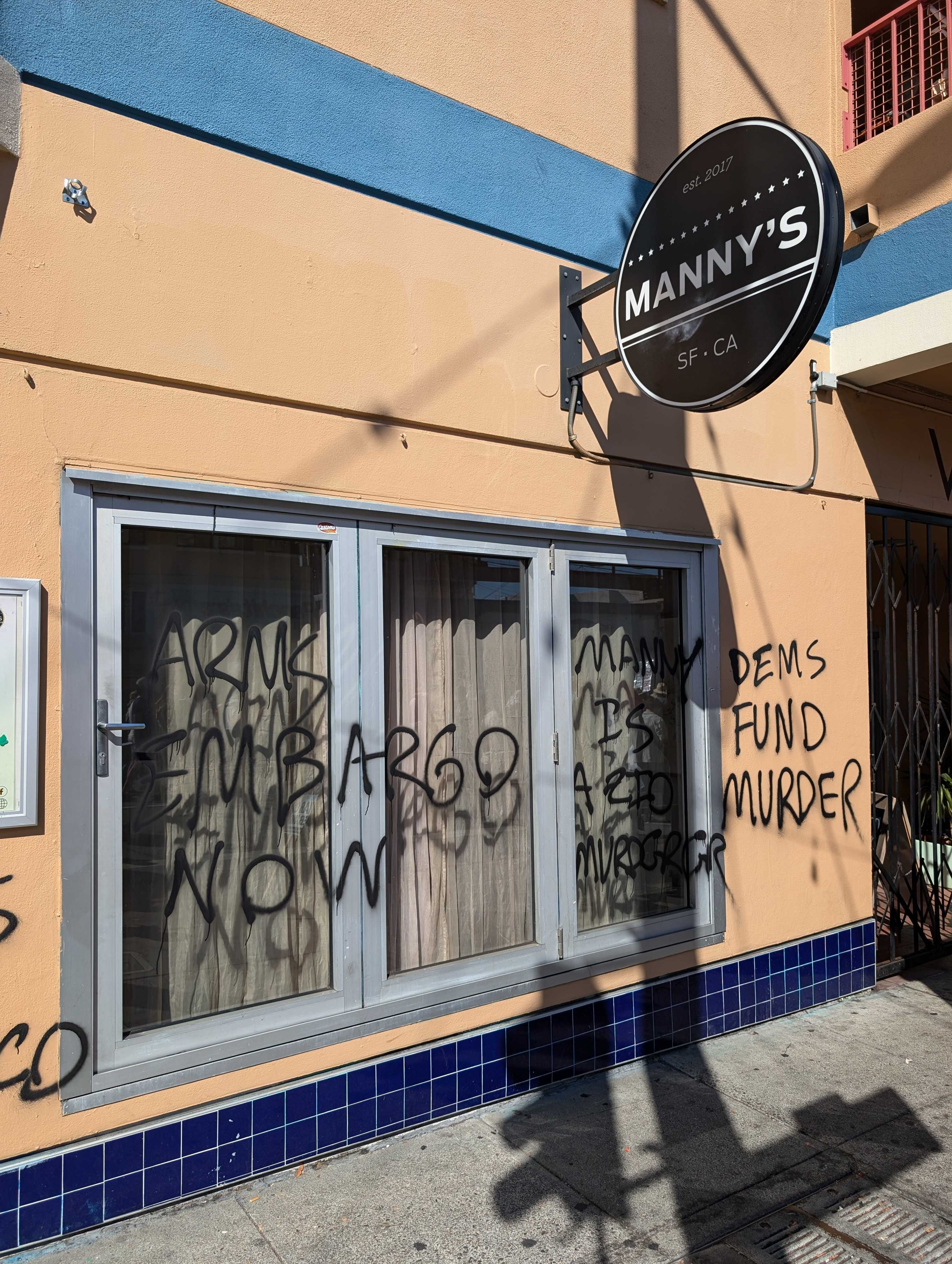 A storefront sign reads &quot;Manny's SF-CA&quot; with graffiti on the windows and wall saying &quot;Arms Embargo Now&quot; and &quot;Dems Fund Murder.&quot;