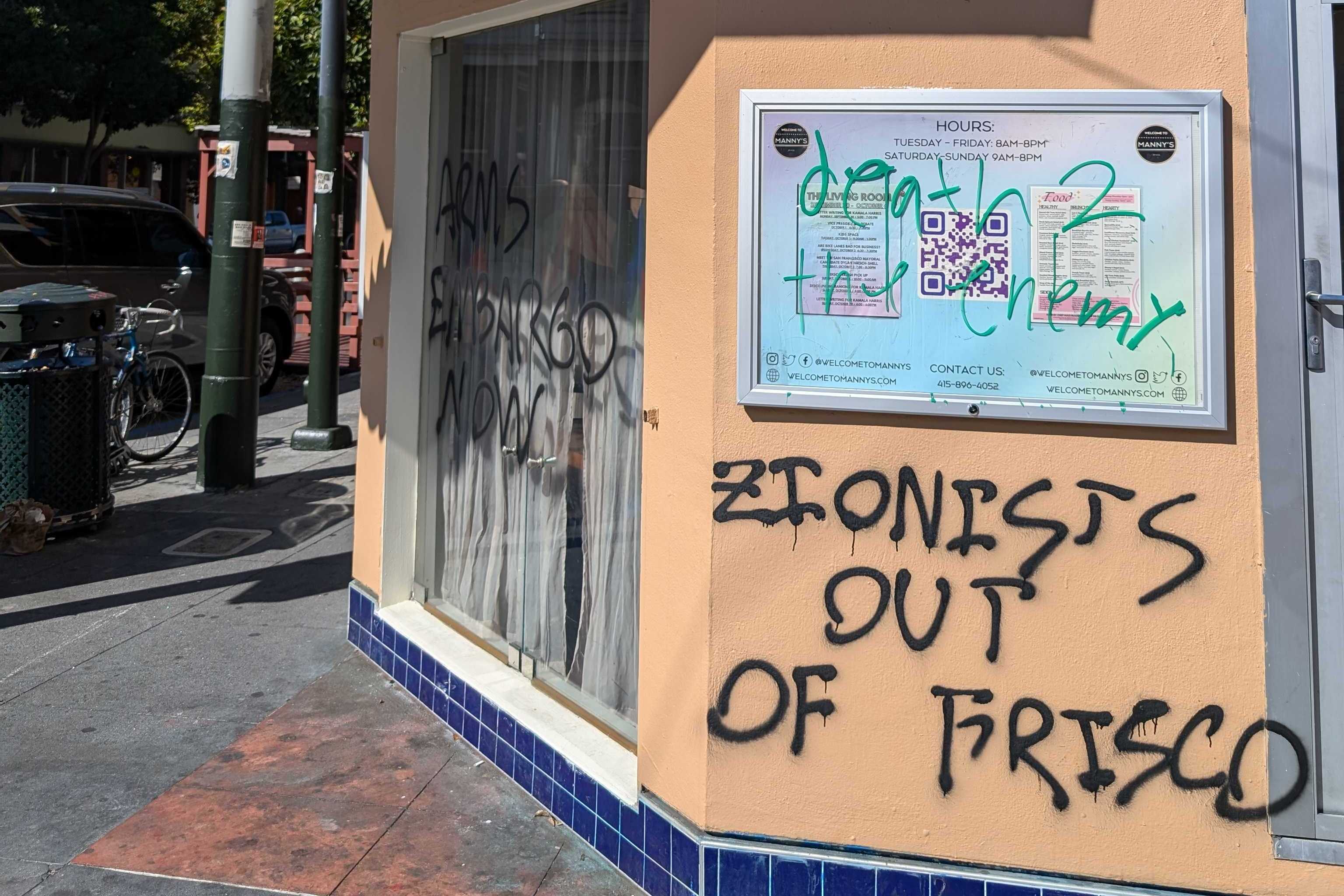 A street corner building shows graffiti reading "ARMS EMBARGO NOW" and "ZIONISTS OUT OF FRISCO" on its walls. There's a notice board and a QR code nearby.