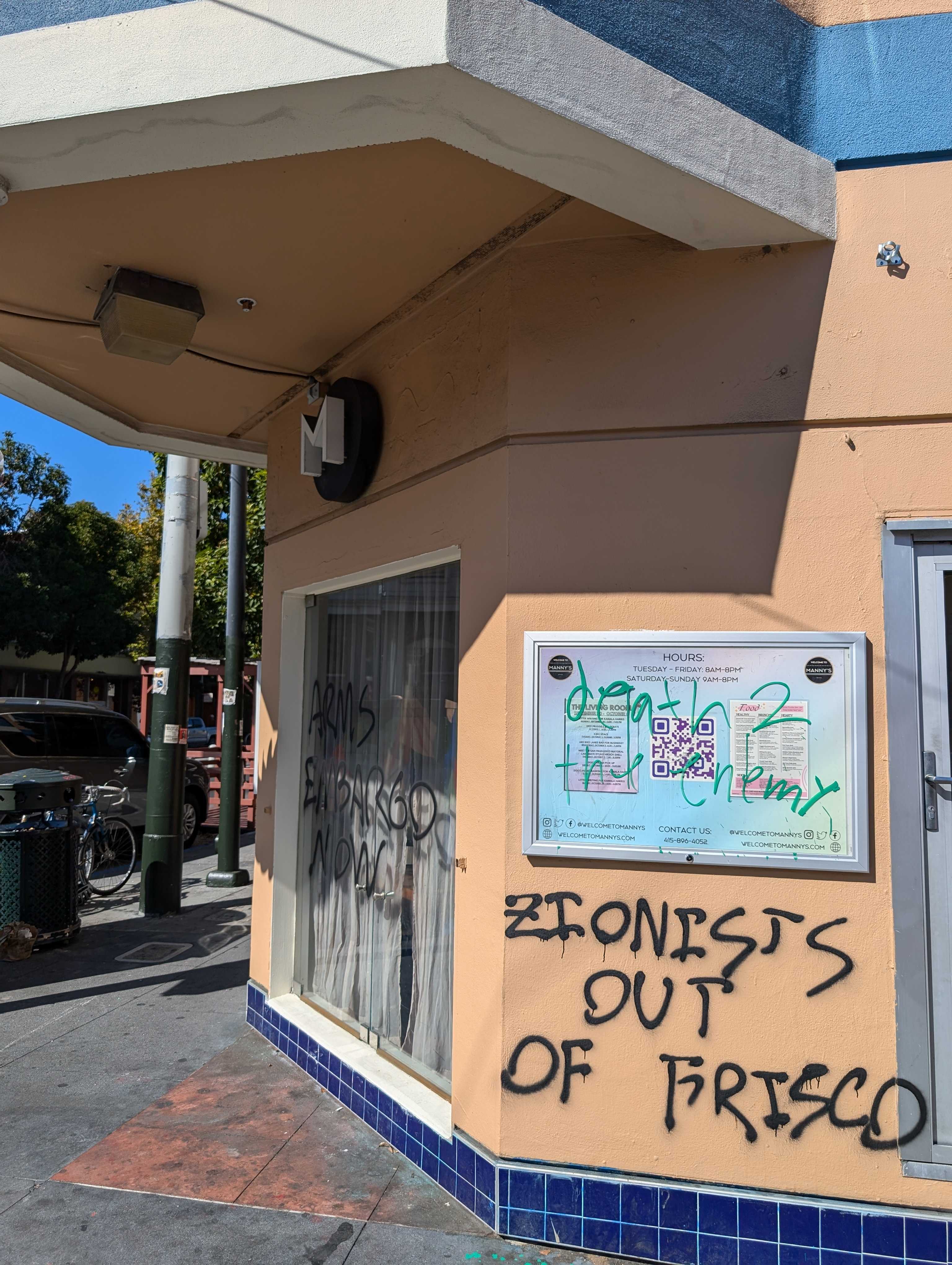 A street corner building shows graffiti reading "ARMS EMBARGO NOW" and "ZIONISTS OUT OF FRISCO" on its walls. There's a notice board and a QR code nearby.