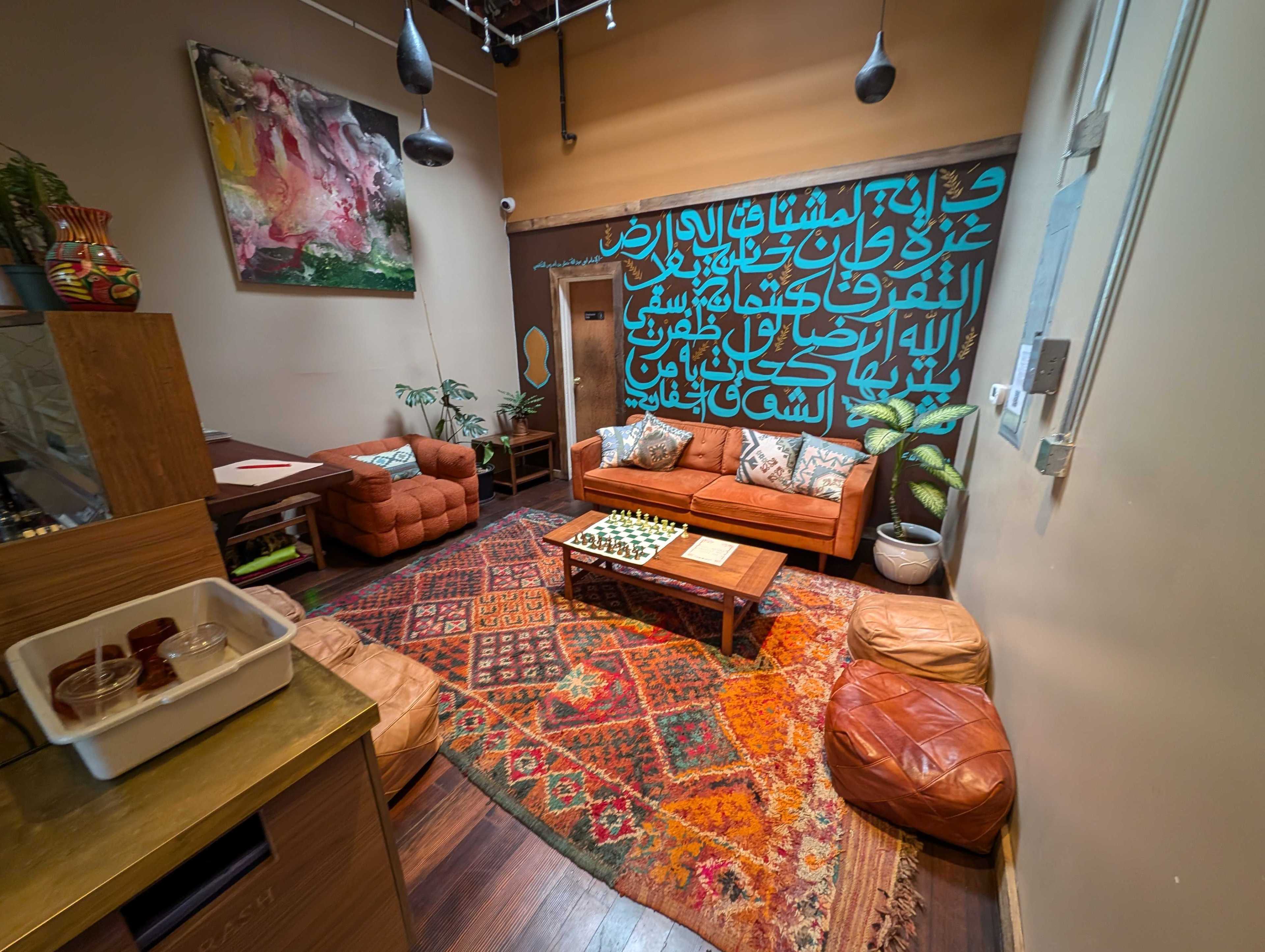 A cozy room features a brown leather sofa, patterned rug, abstract art, large wall script, plants, and a chessboard on a wooden table. Warm lighting enhances the ambiance.