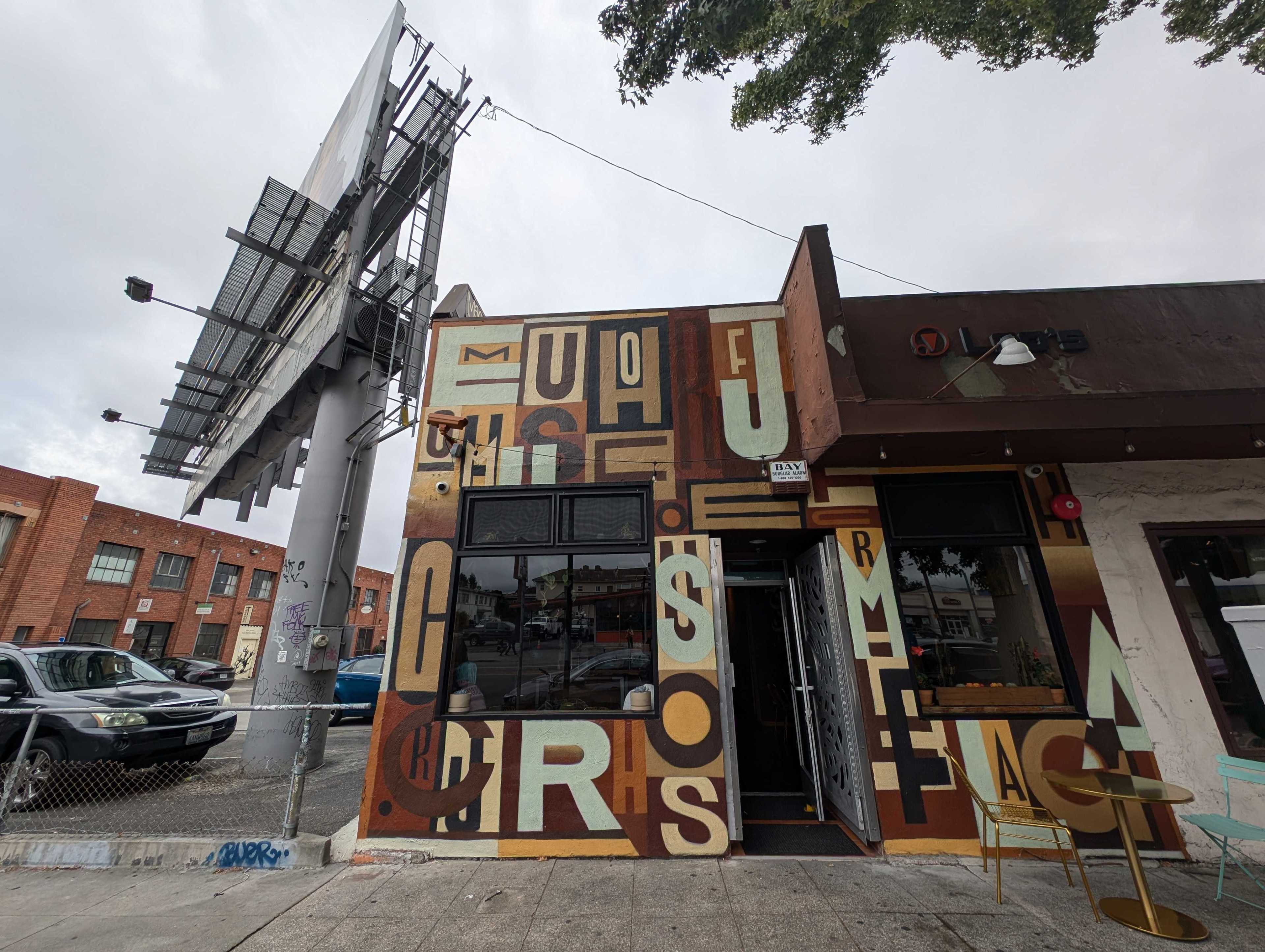 A cafe exterior with eclectic mural art displaying large letters in various colors. A metallic chair and round table sit outside, and a large billboard towers nearby.