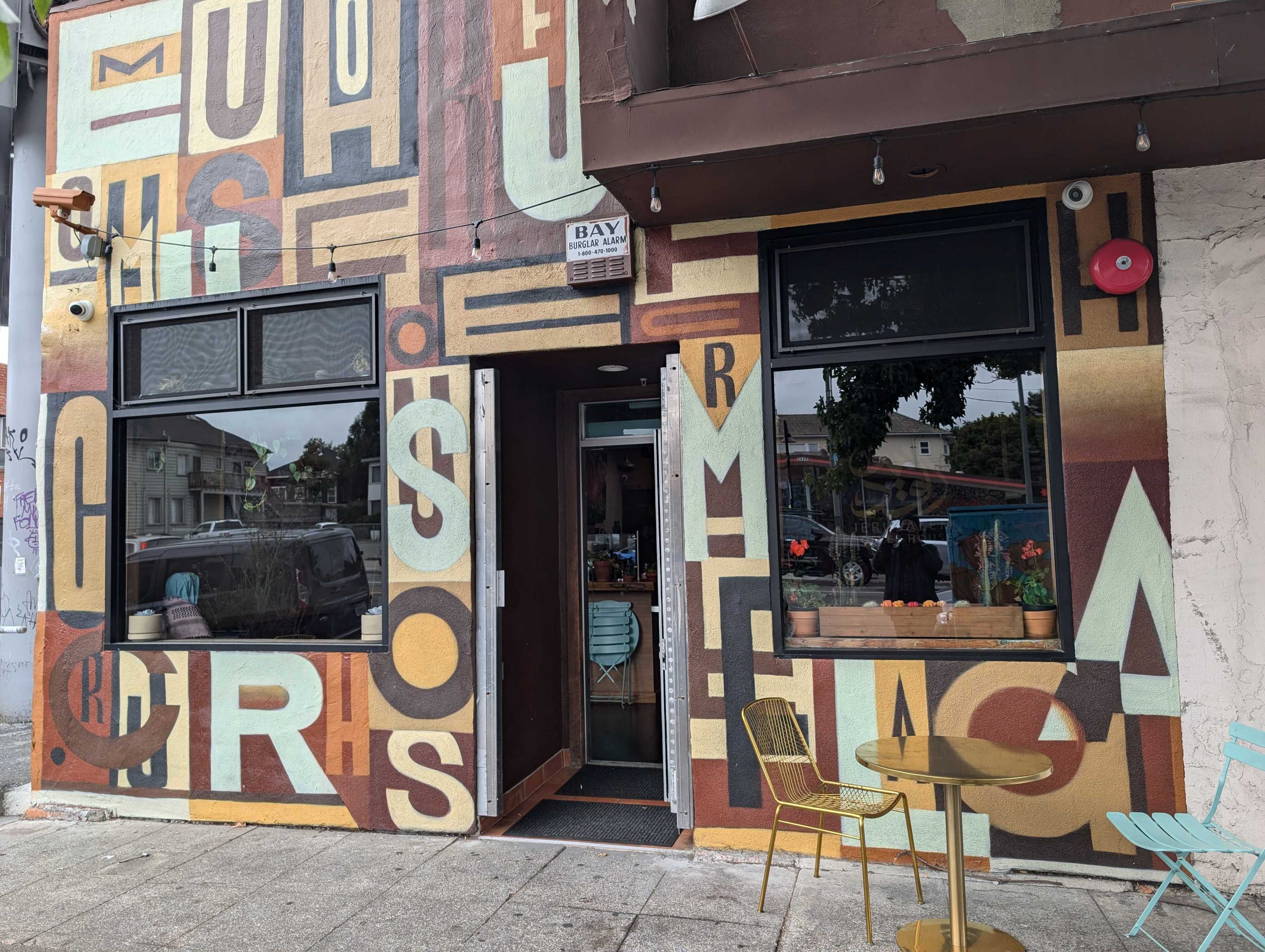 The image shows a colorful cafe exterior with large, overlapping letters painted on the walls. There are two windows and an open door, plus a small outdoor table with chairs.