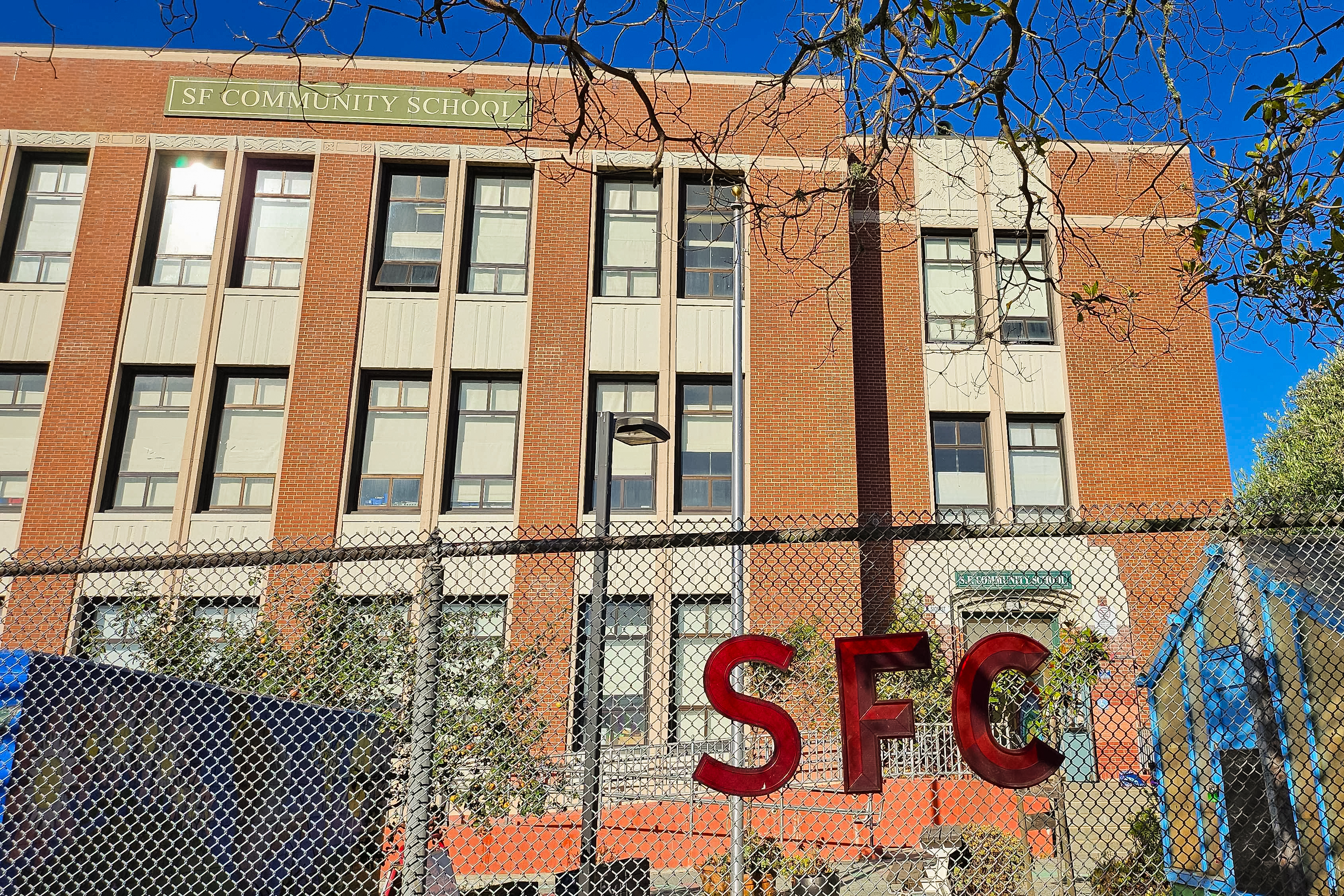 A brick building with a "SF Community School" sign is visible. In front, a chain-link fence features red letters spelling "SFC." Trees and a bright blue sky surround it.